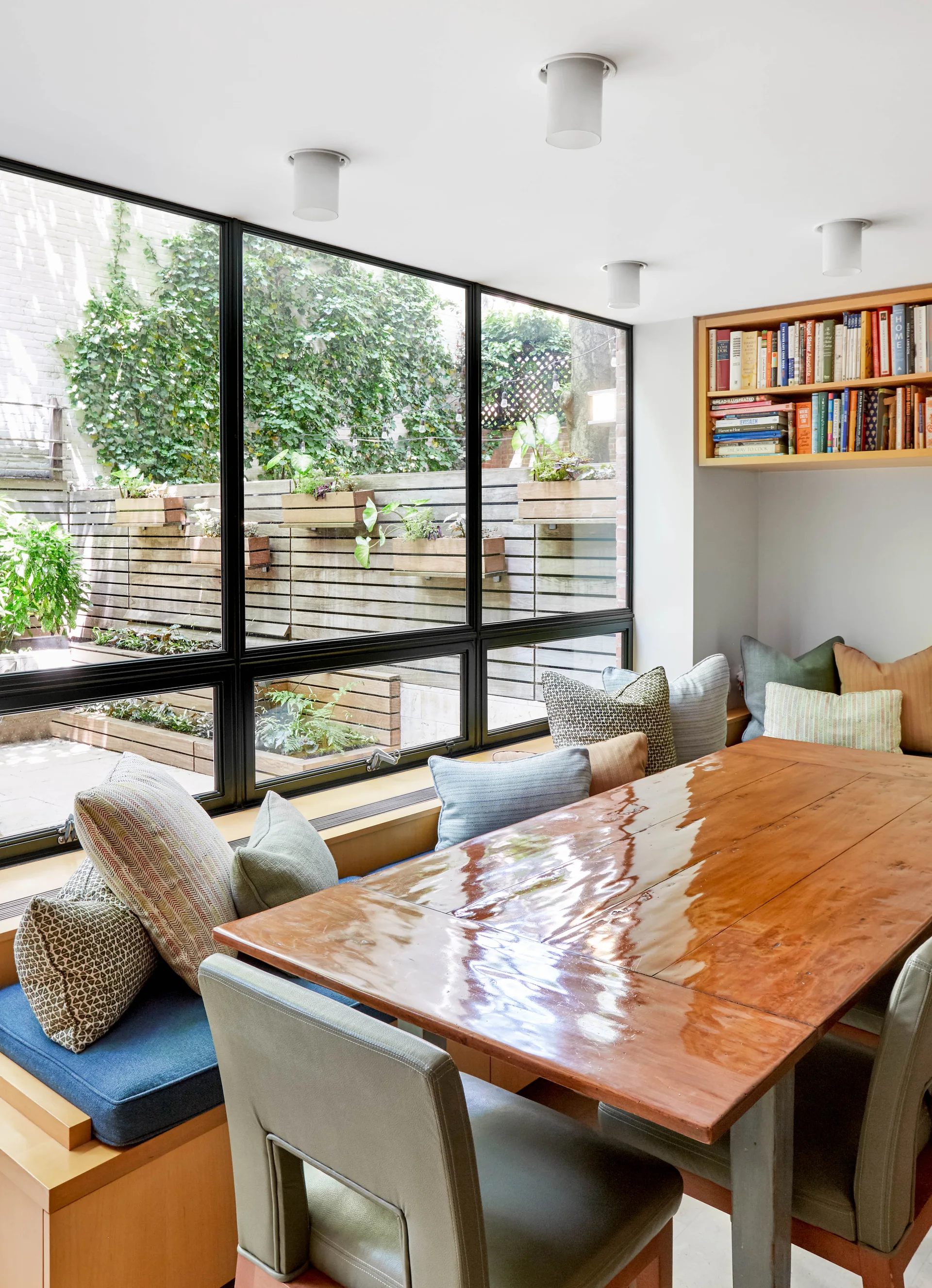 A dining room featuring a glossy wood table, upholstered chairs, a built-in window bench, and large black-framed windows looking out at a garden patio.