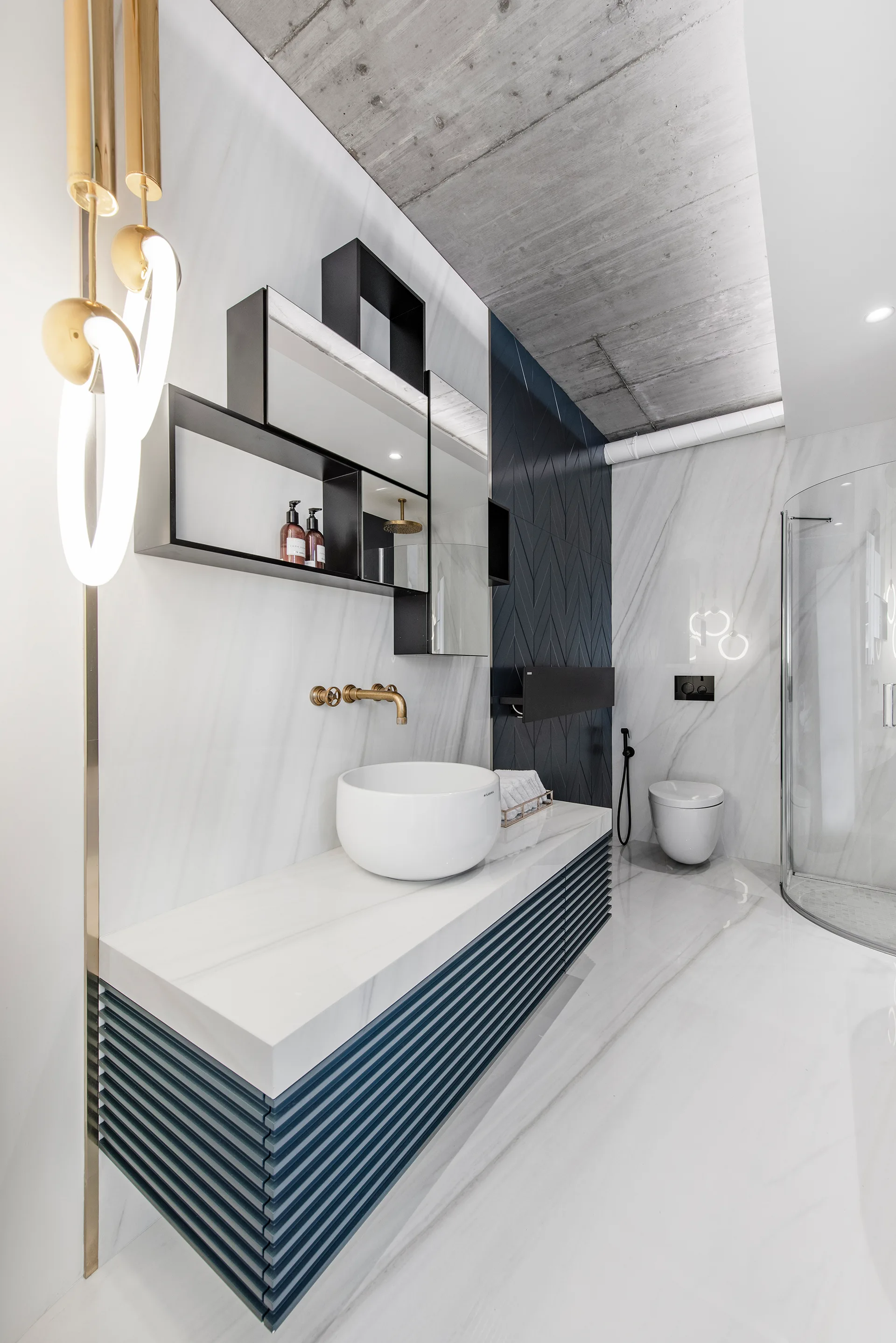 A modern bathroom features a white marble vanity with a navy slatted base, a round vessel sink, brushed gold fixtures, and an exposed concrete ceiling.