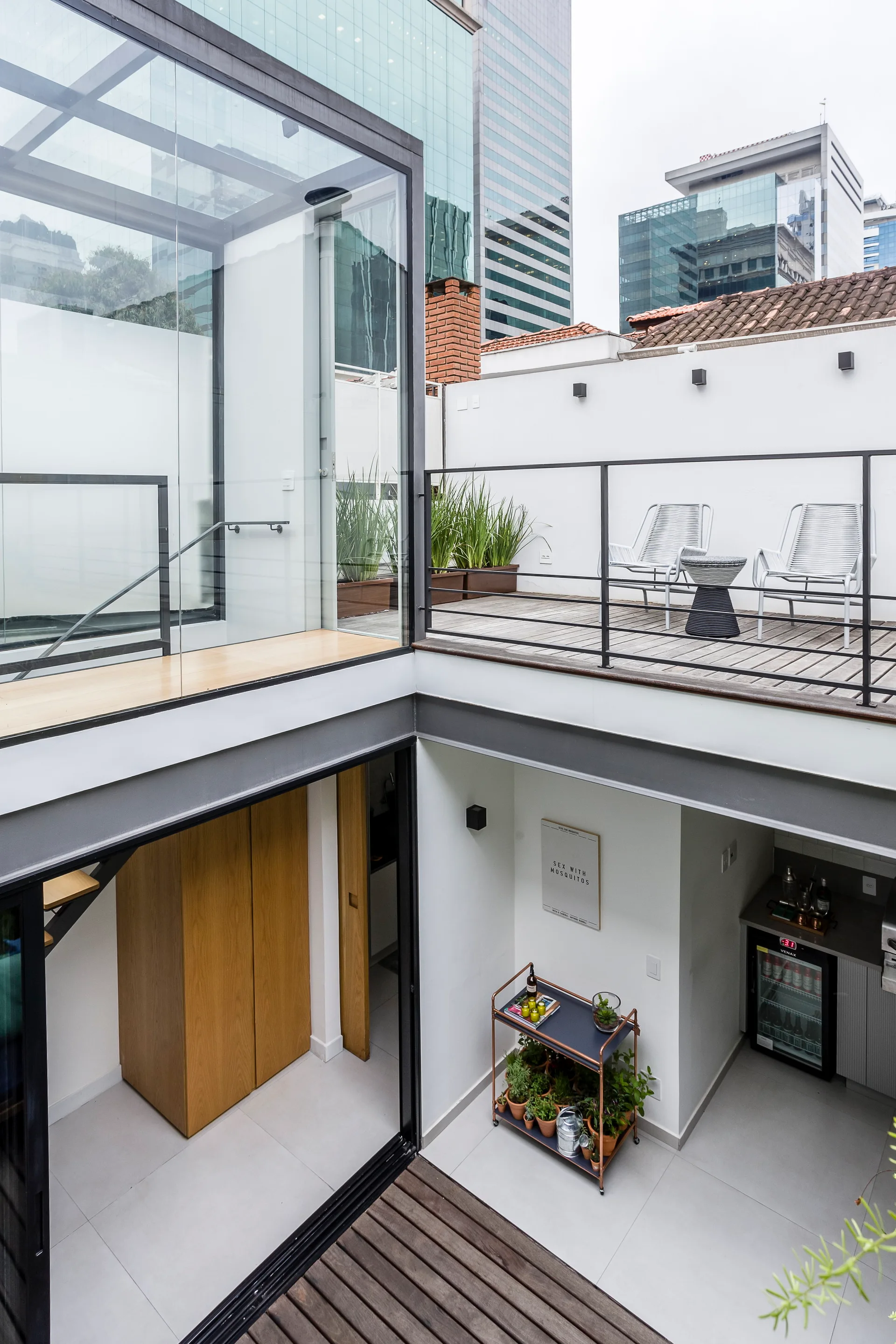 A modern multi-level outdoor terrace features a glass-enclosed staircase, an upper deck with two chairs, and a lower level with a kitchenette.