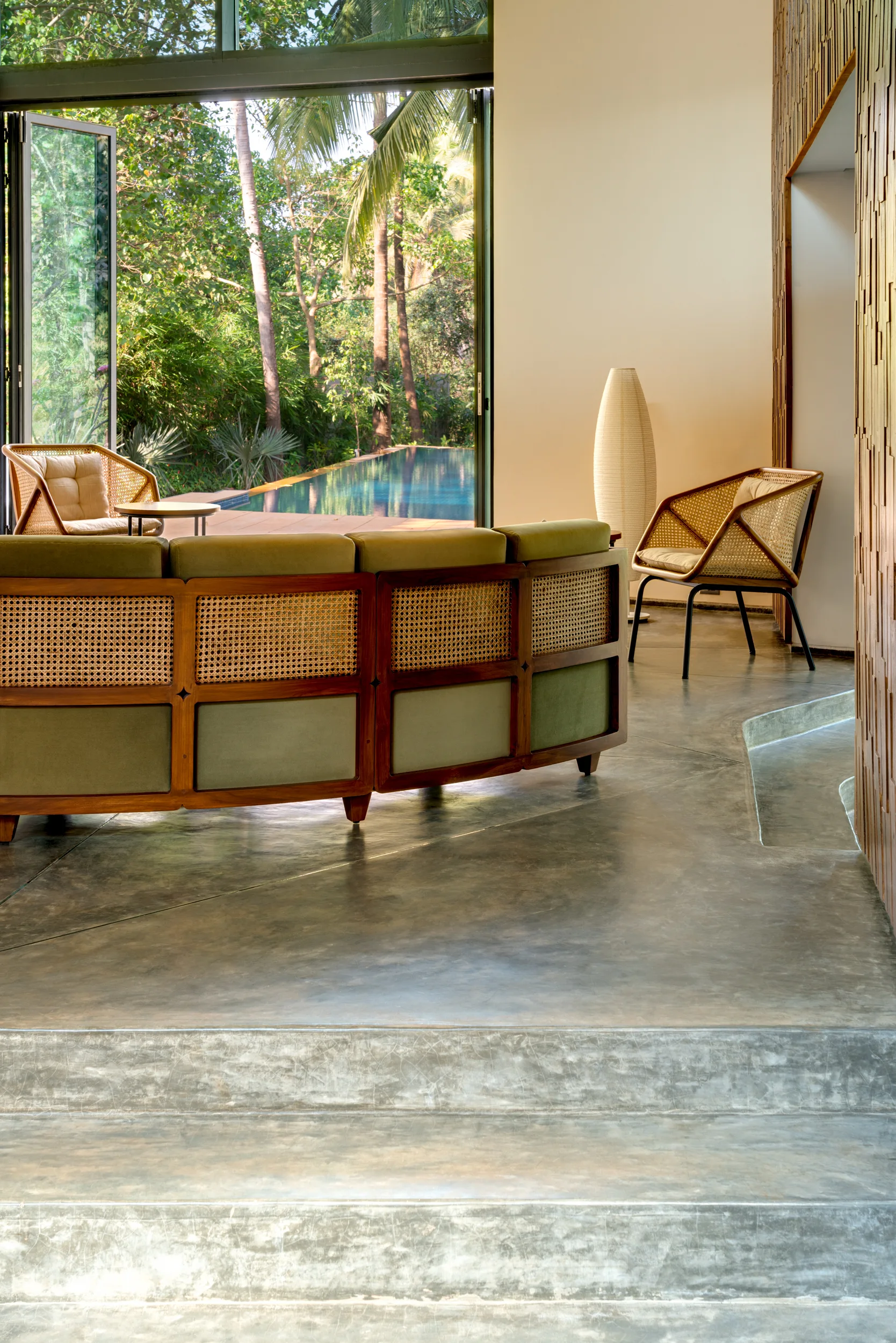 A tropical living room features a curved wood and cane sofa, grey concrete floors, and wide open doors leading to an outdoor swimming pool.