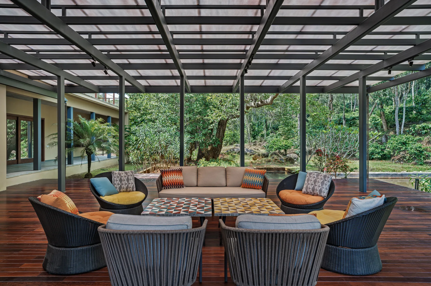A covered outdoor patio features a beige sofa, dark woven armchairs, and patterned coffee tables on polished dark wood decking.