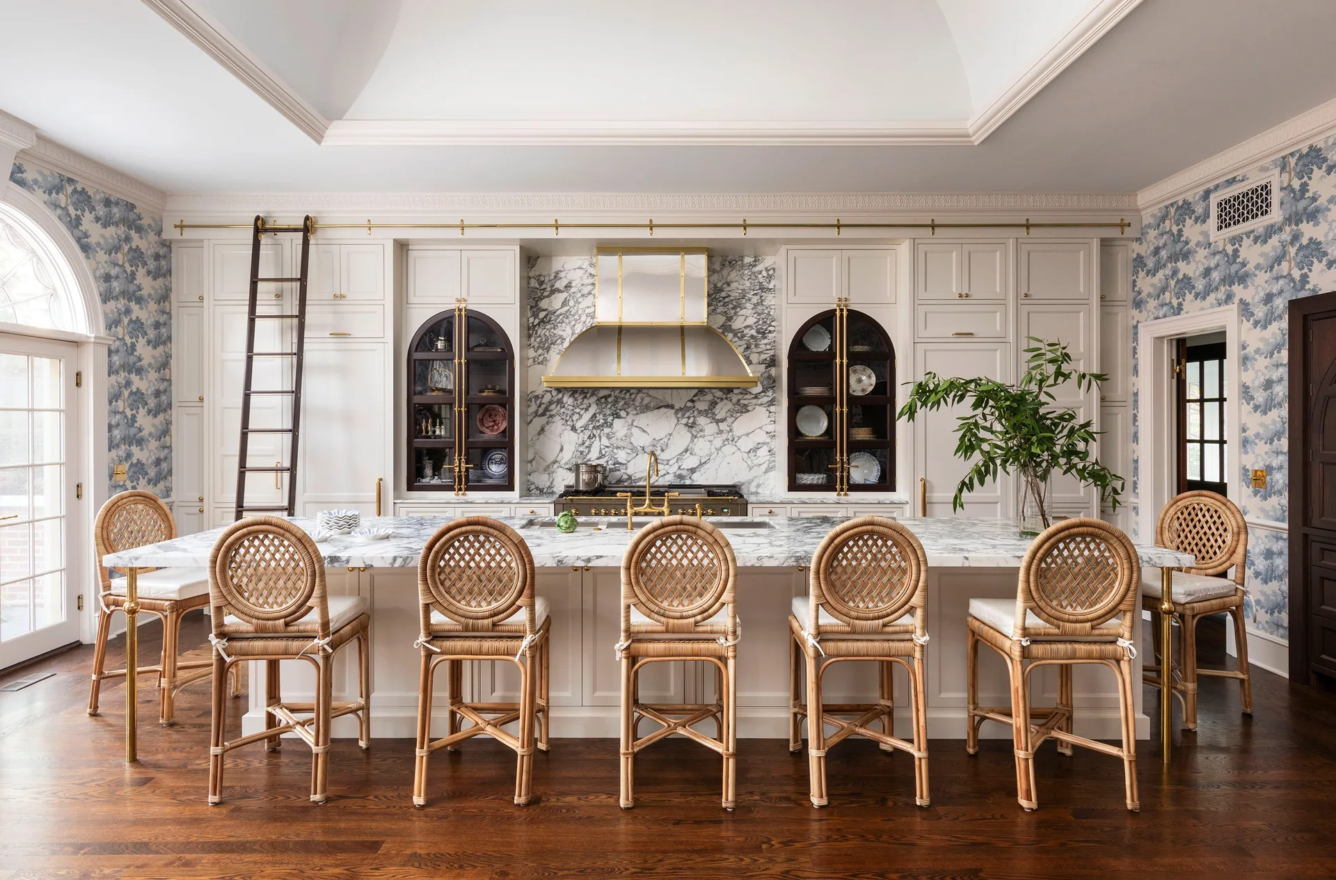 A spacious white kitchen features a large marble-topped island lined with six rattan bar stools beneath a white vaulted ceiling.
