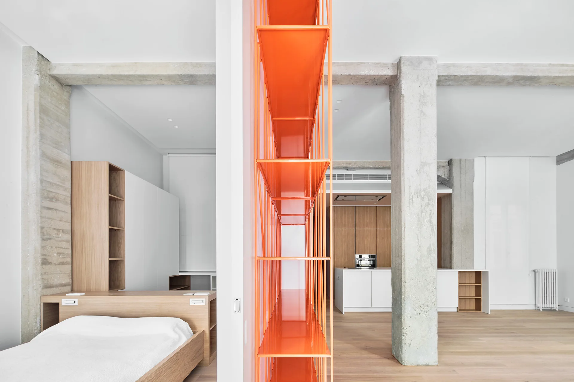 An open-plan room features a bright orange steel shelving unit acting as a divider between a bedroom space and a kitchen with concrete columns.