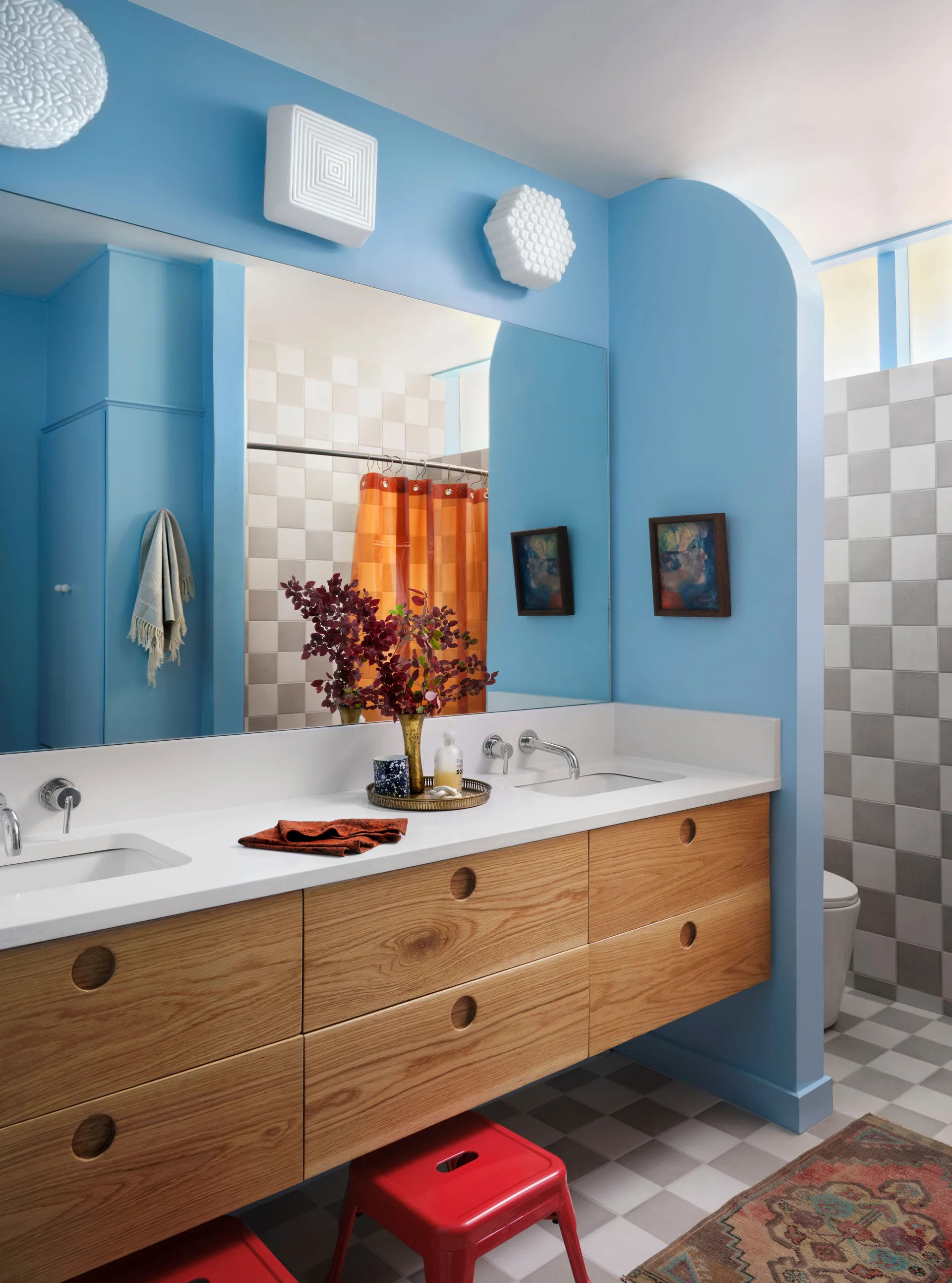 A bathroom features bright blue walls, a floating oak double vanity with round recessed pulls, a large mirror, and grey and white checkerboard floor tiles.