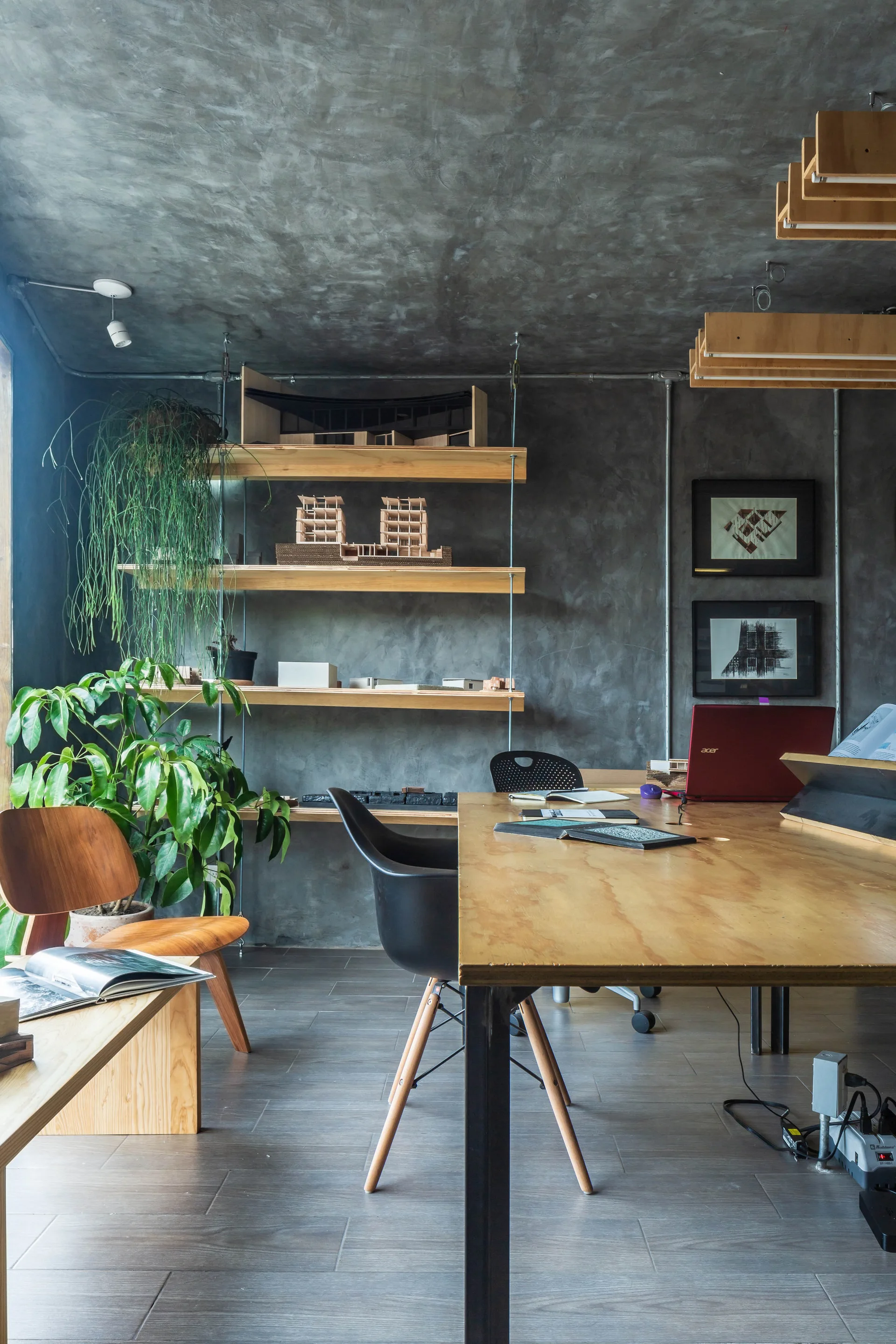 An industrial office space features a wood desk, a black chair, open plywood shelves with architectural models, and raw concrete walls.