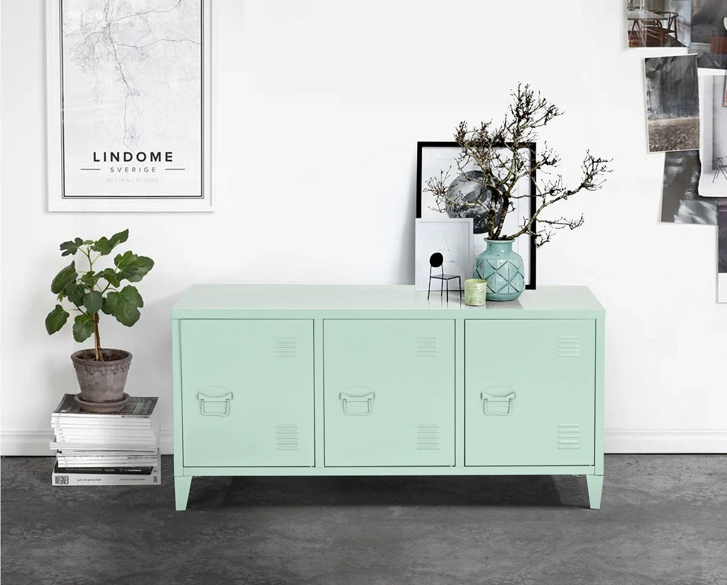 A three-door mint green metal locker cabinet sits against a white wall next to a stacked pile of books with a small potted plant.