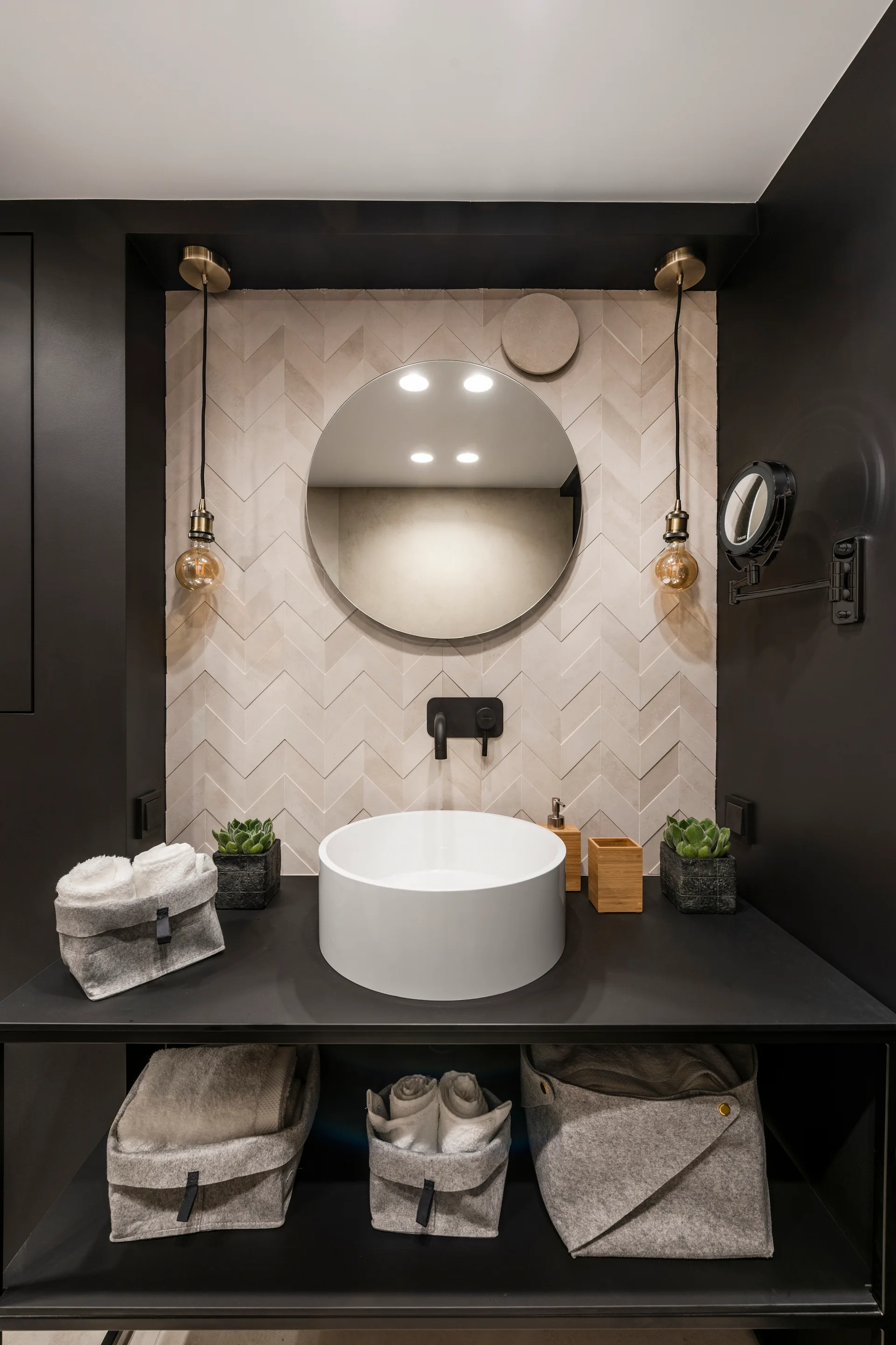 A minimalist bathroom features a black console vanity with a white vessel sink, a round mirror, and a beige chevron-tiled accent wall.