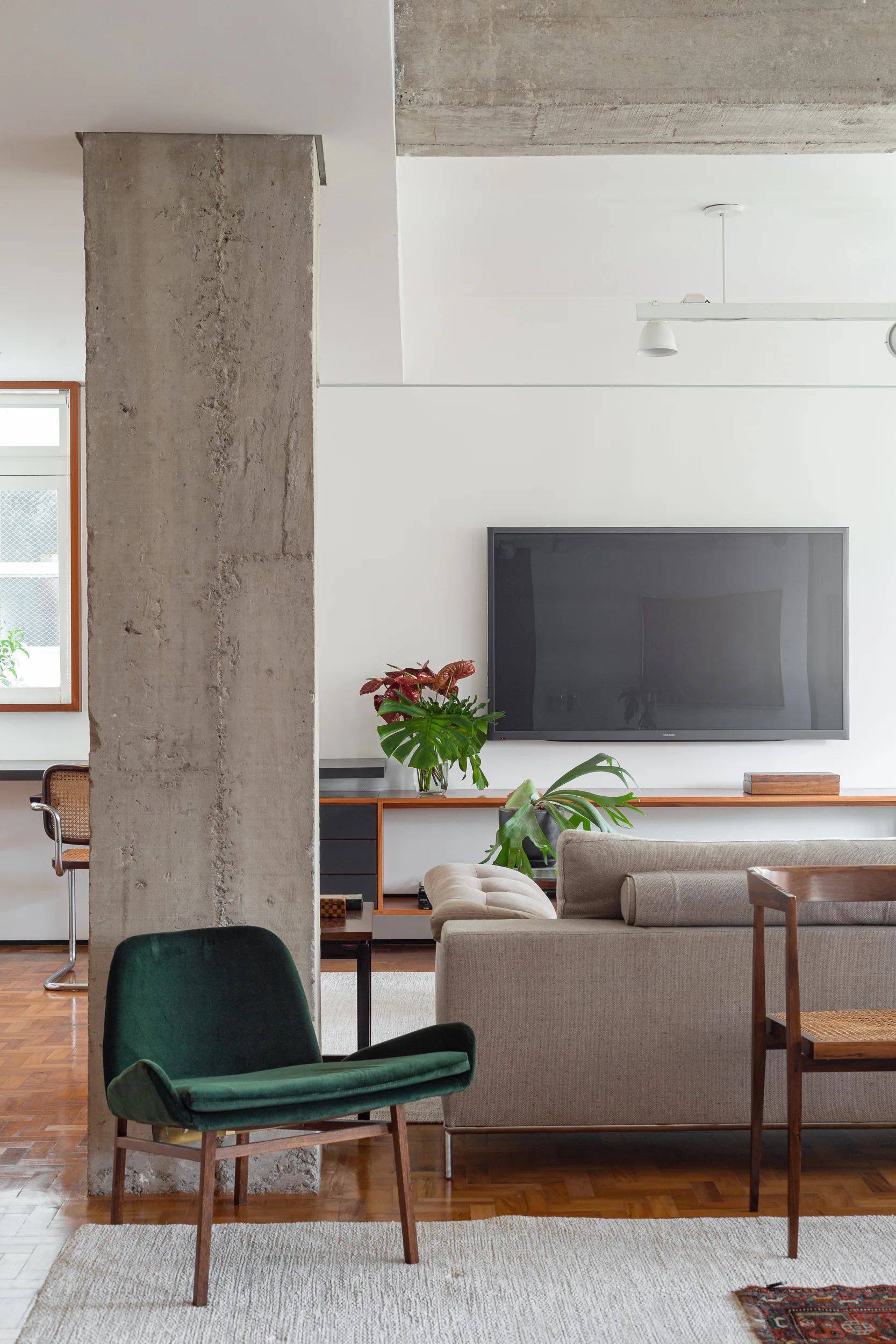 A modern living room features a green velvet armchair, neutral sofa, and wall-mounted television next to a prominent raw concrete column.