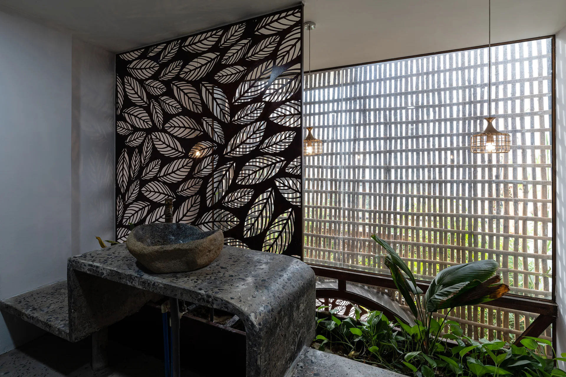A grey stone bathroom vanity with a rough stone basin is set against a metal leaf-pattern screen and a window covered in vertical bamboo slats.