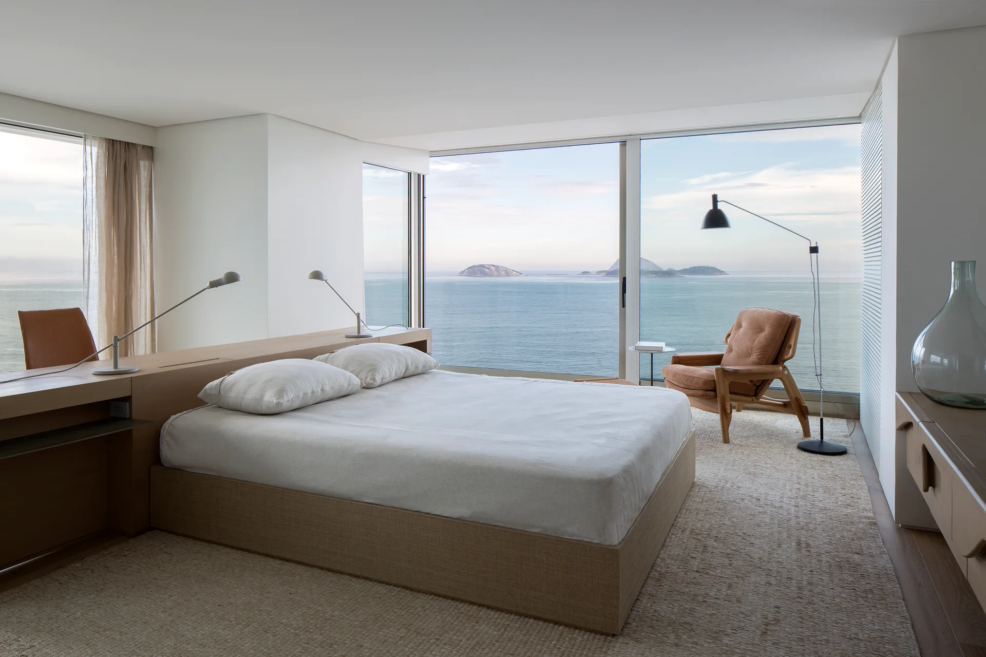 A minimalist bedroom features a low bed with an integrated wooden headboard desk, set before floor-to-ceiling windows overlooking the ocean.