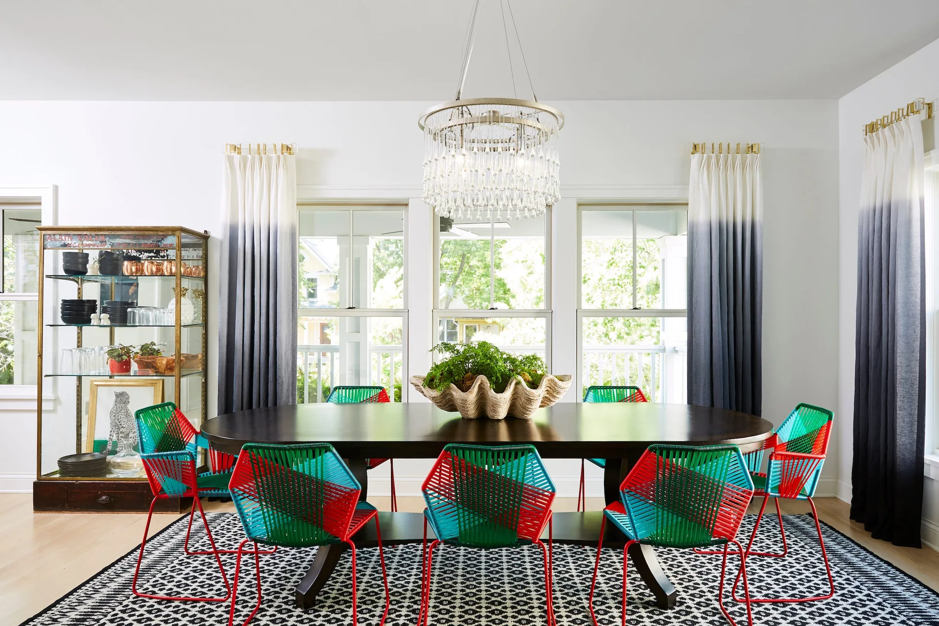 An eclectic dining room features a dark oval wood table surrounded by colorful woven chairs, ombre curtains, and a tiered crystal chandelier.