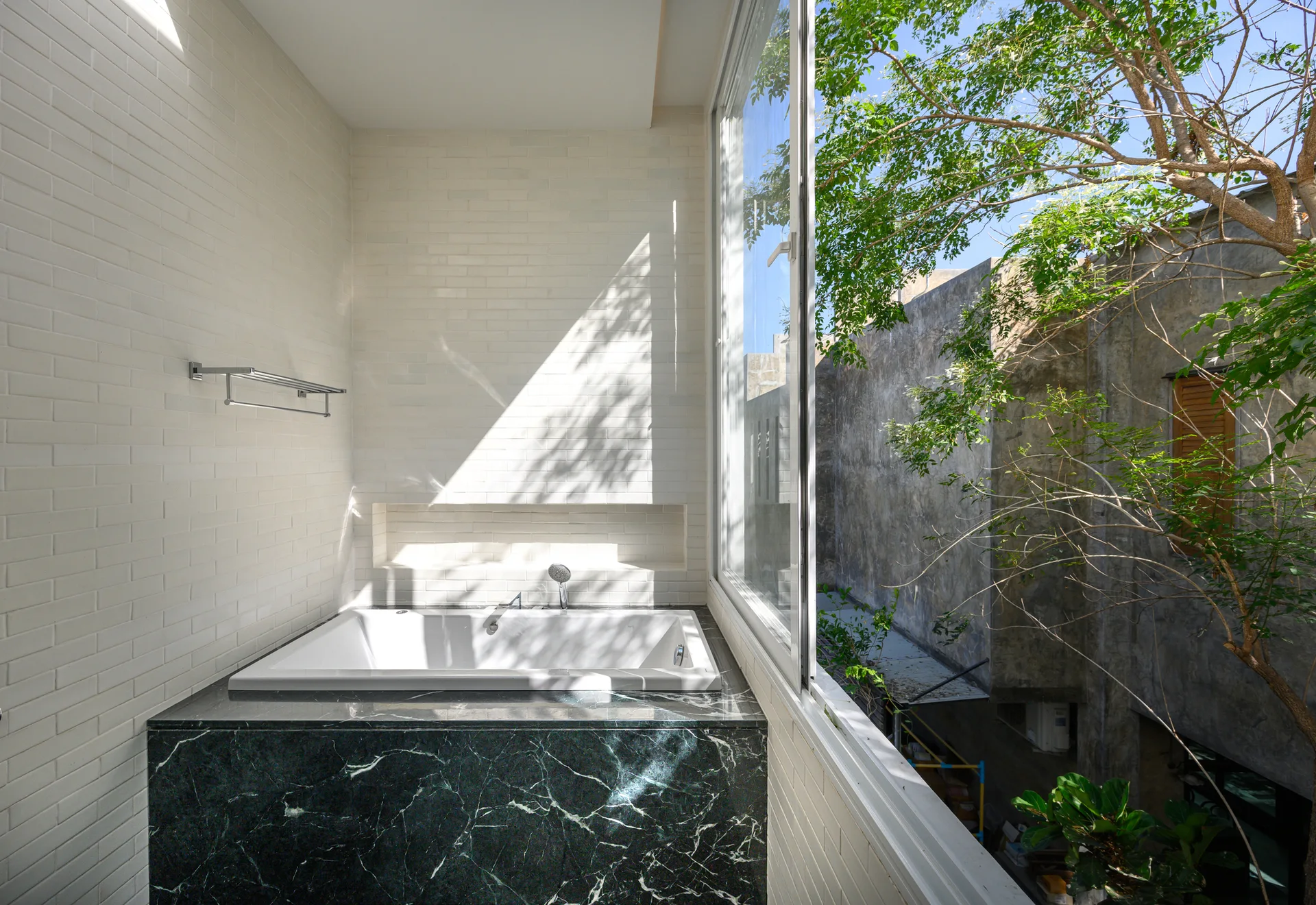 A white bathtub encased in dark green veined marble sits against white brick walls next to a large sliding glass window looking out at trees.