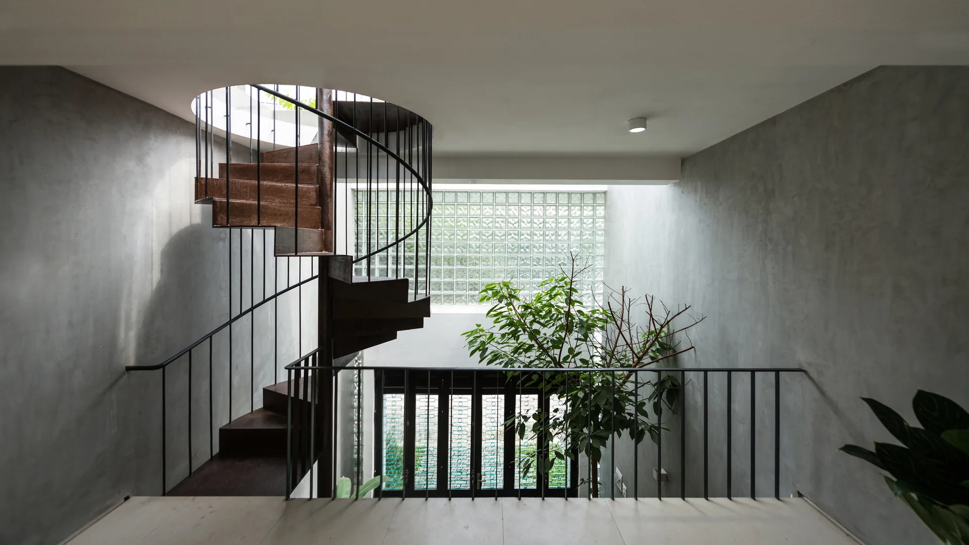 A dark metal spiral staircase stands in a minimalist interior featuring smooth concrete walls, a black metal railing, and a glass block window.