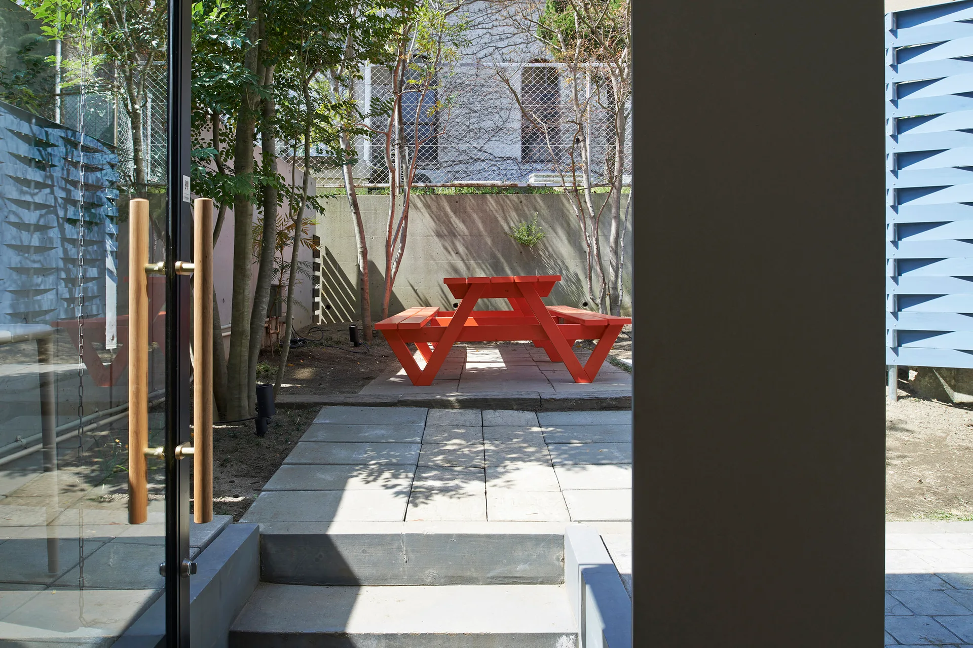 A bright orange wooden picnic table sits on a grey stone paved patio surrounded by slender trees and a concrete wall.