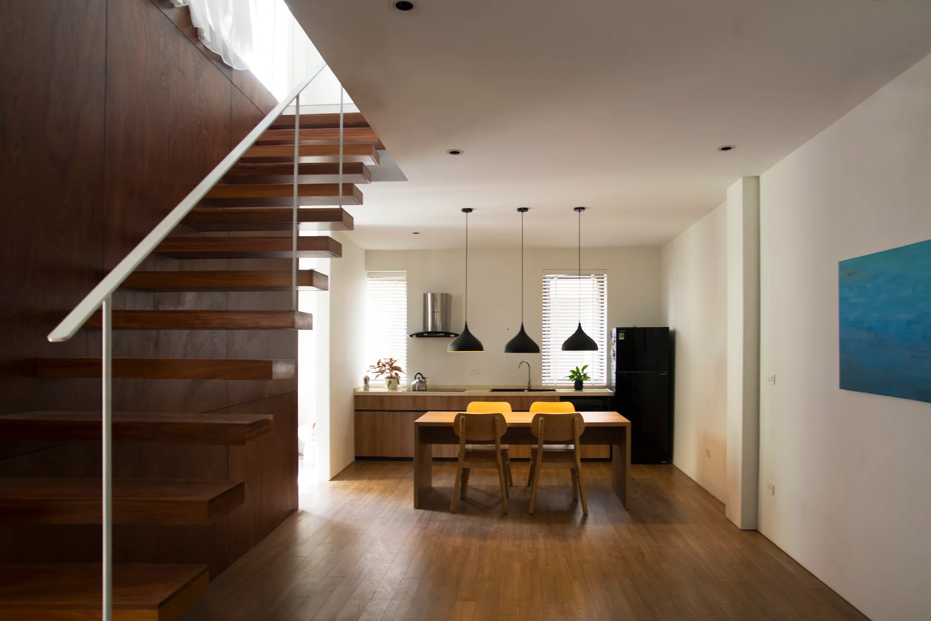 A minimalist open-plan kitchen and dining area featuring a floating wooden staircase, a wood dining set, and three black pendant lights.