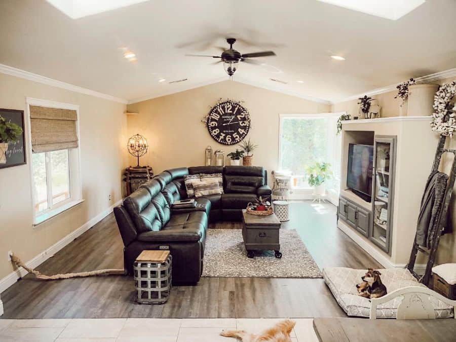 A spacious living room with neutral beige walls, a dark leather sectional sofa, a large round wall clock, and a gray built-in media center.