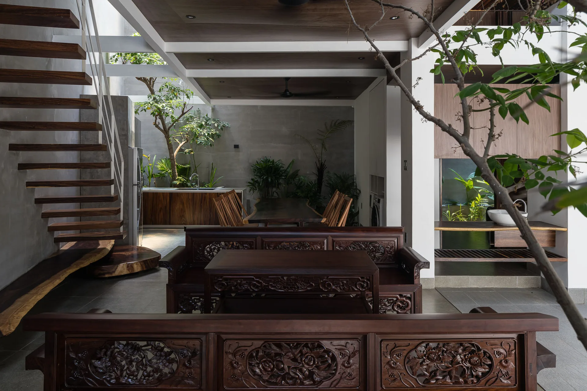 A contemporary open-concept living area features a carved dark wood sofa, a floating wooden staircase, and an open courtyard with plants.