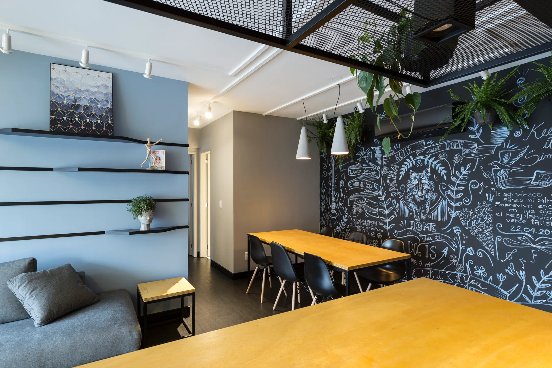 A modern dining area features a wooden table with black chairs, a blue accent wall with floating shelves, and a large hand-drawn chalkboard wall.