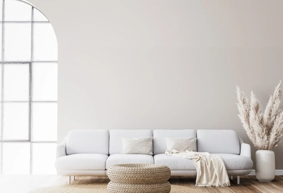 A minimalist living room features a white sofa, a woven wicker ottoman, a fluted vase with pampas grass, and a large arched window.