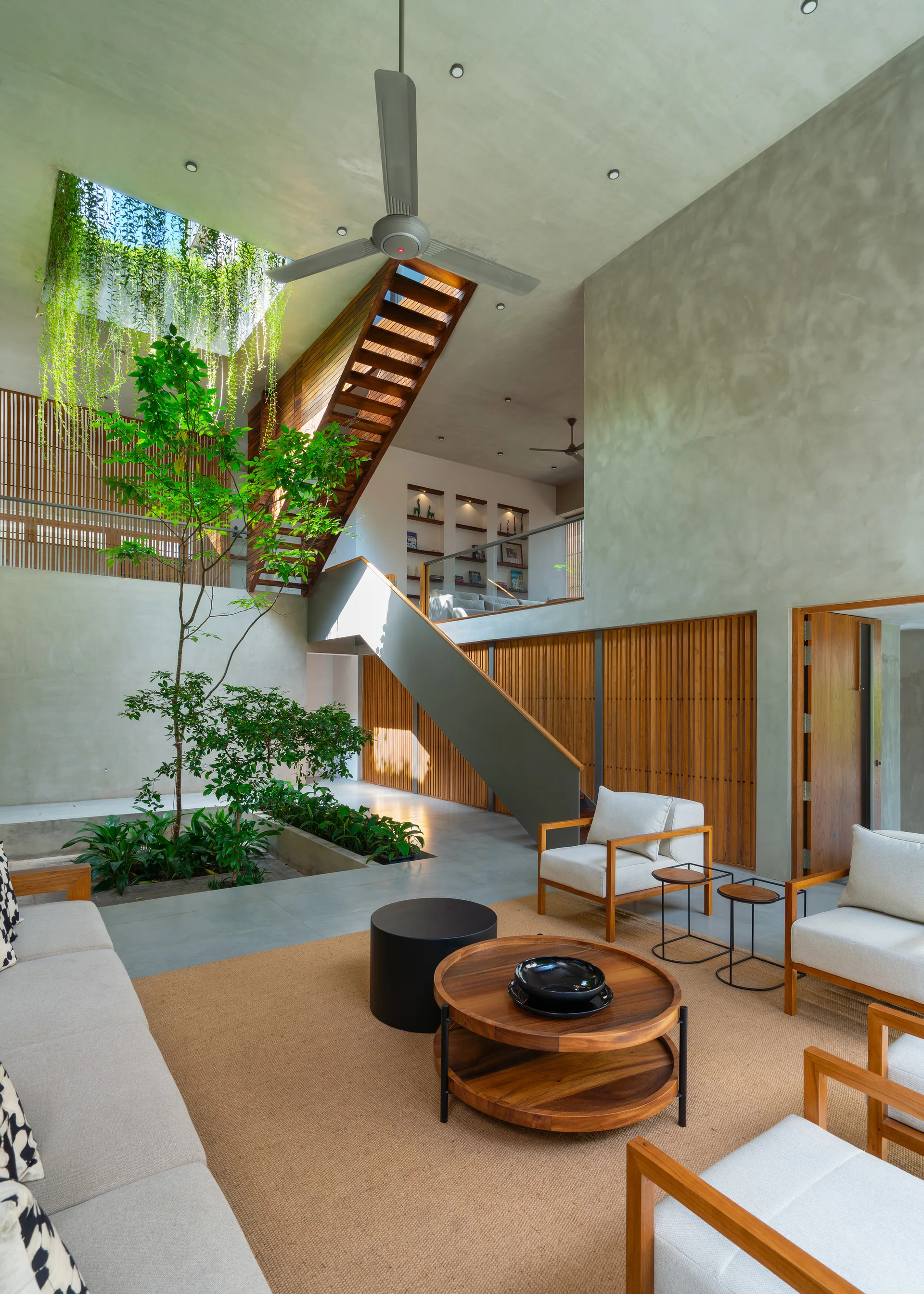 A double-height concrete living room features upholstered armchairs, a central skylit indoor garden with a growing tree, and a wooden staircase.