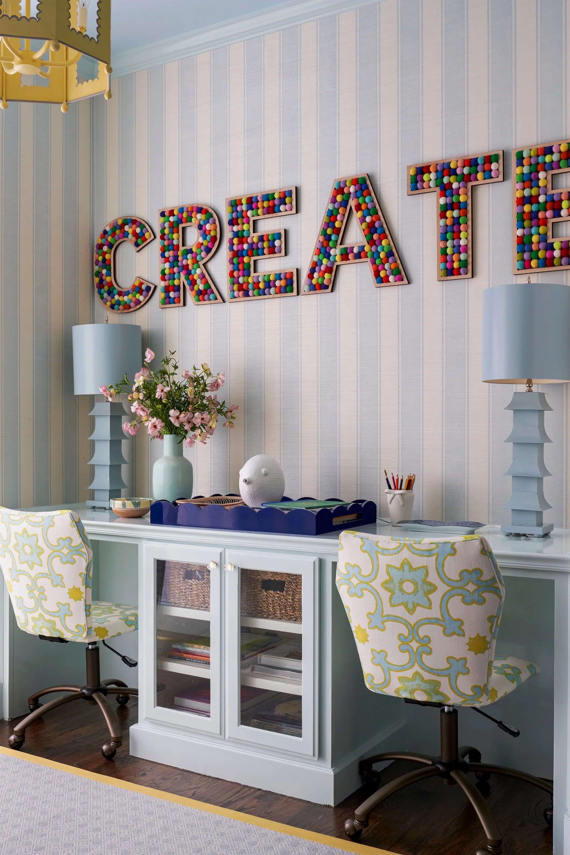 A shared light blue built-in desk with two patterned chairs is set against striped wallpaper beneath multi-colored pom-pom letters spelling CREATE.