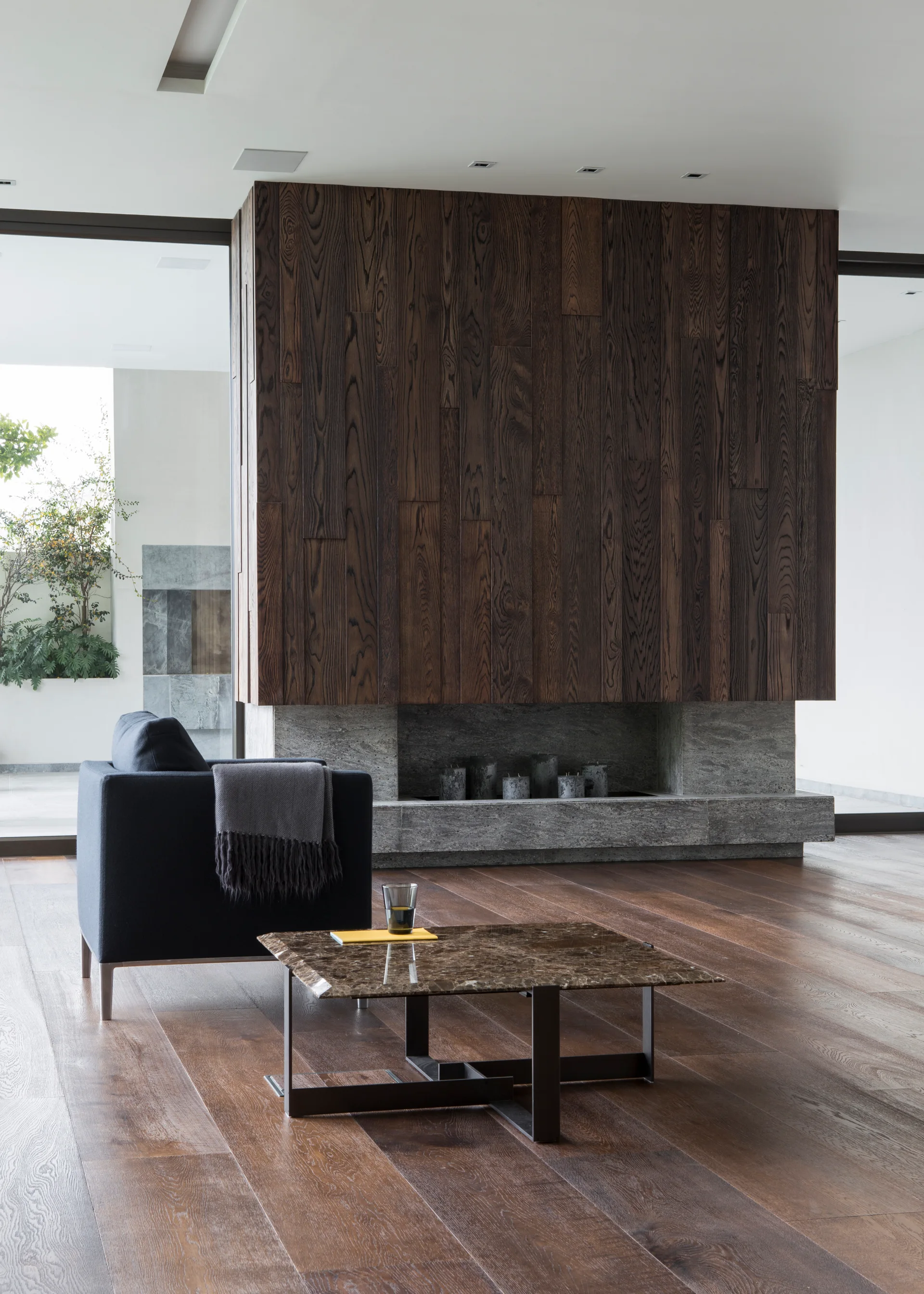 A modern living room features a dark vertical wood paneled fireplace with a stone base, a dark armchair, and a marble coffee table on wide-plank floors.