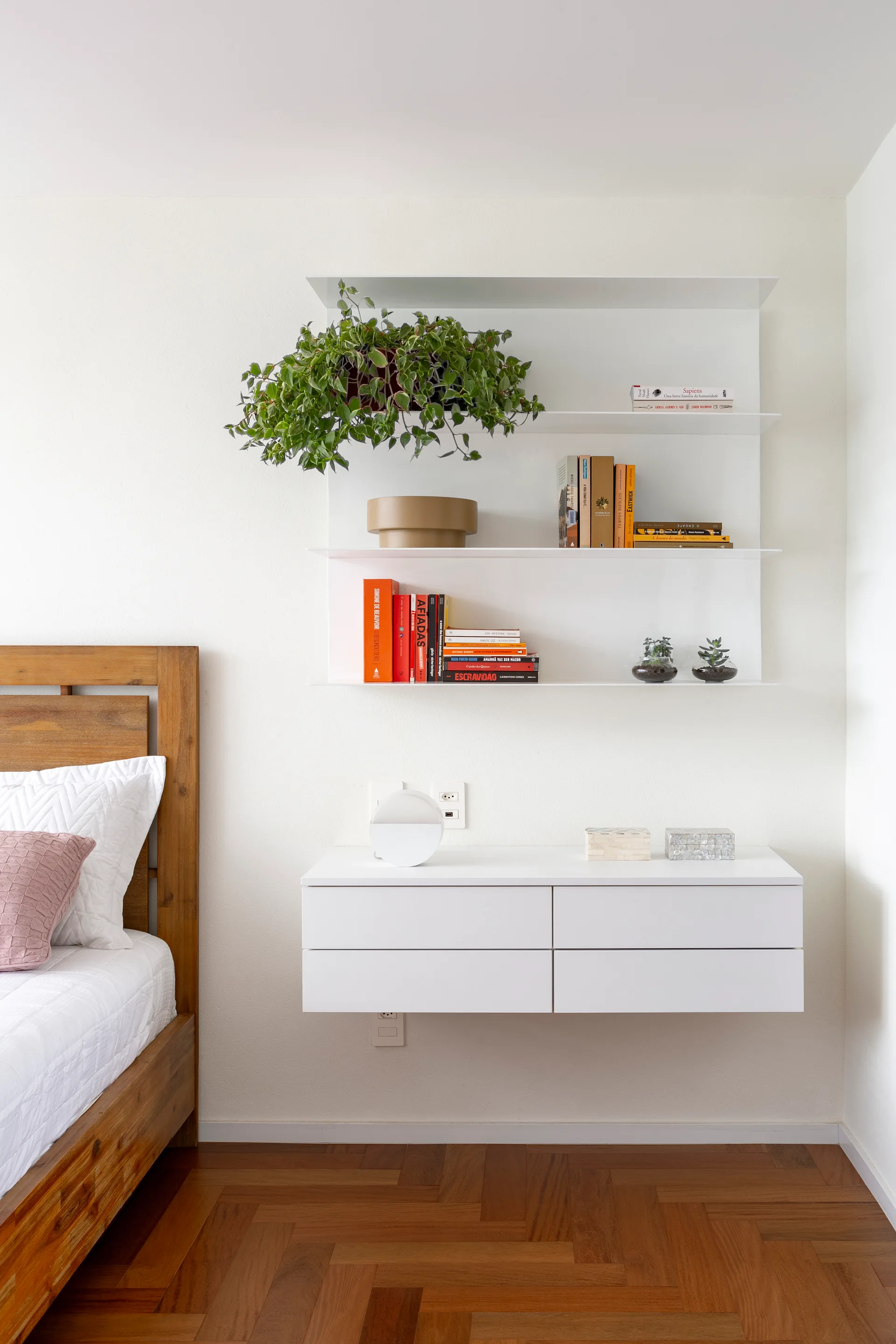 A minimalist bedroom features a wooden bed frame next to a white floating console and wall-mounted shelving unit on a parquet floor.