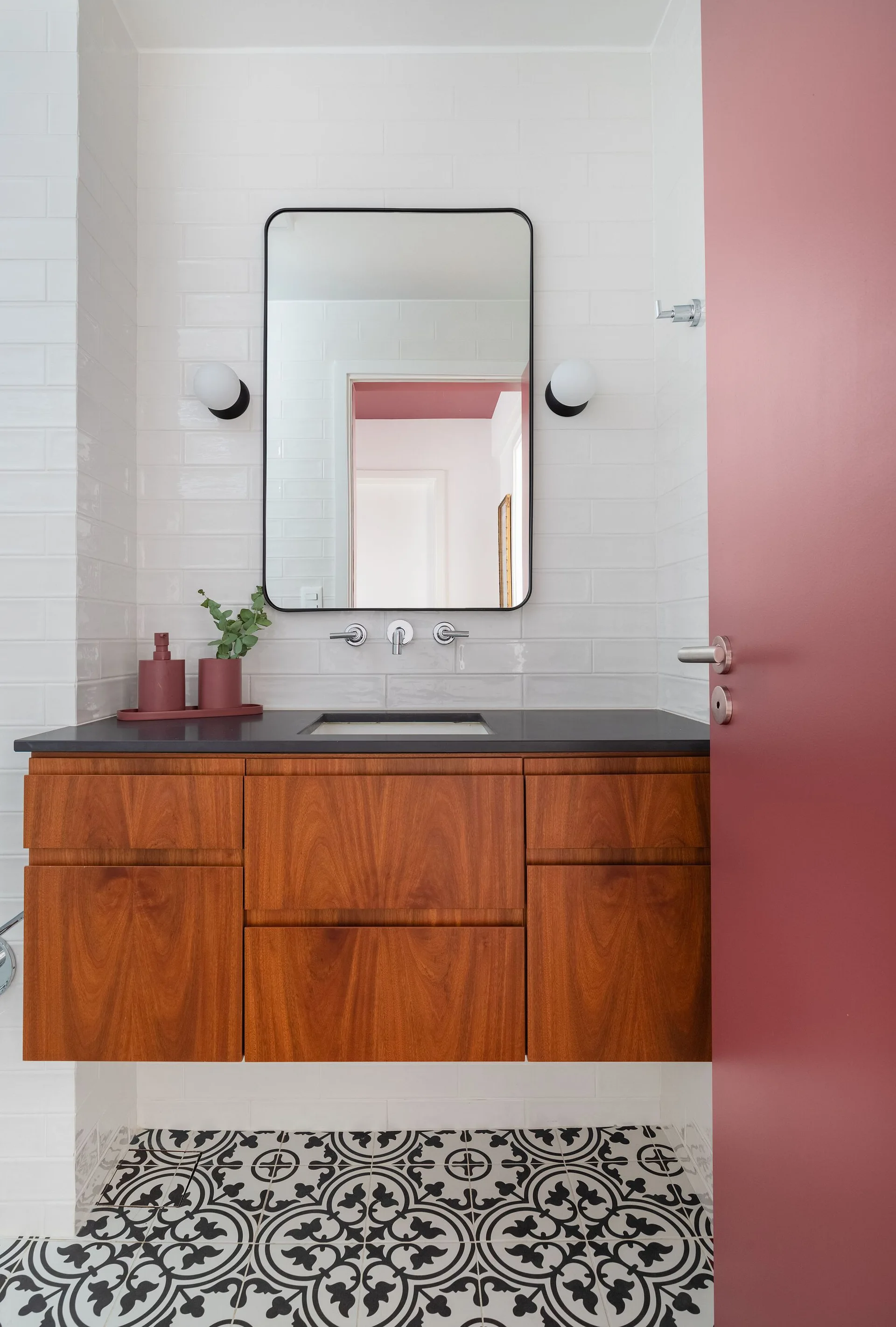 A modern bathroom features a floating wood vanity, white subway tile walls, a black-framed mirror, and a bold pink door.