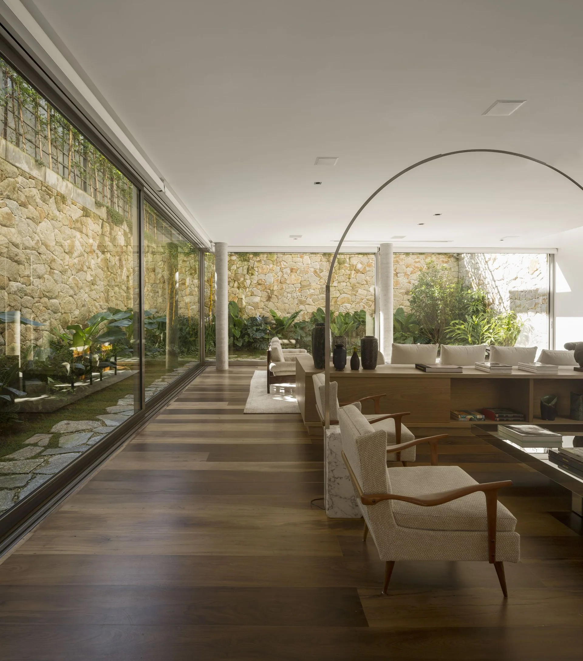 A spacious living room features wide-plank wood floors, mid-century modern armchairs, and floor-to-ceiling glass walls facing a stone-walled garden.