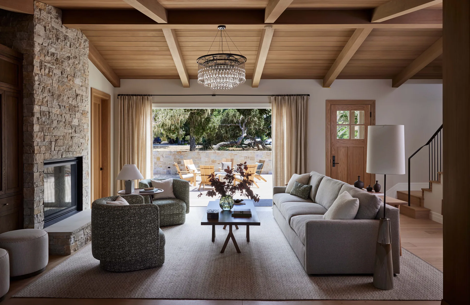 A living room features a textured stone fireplace, wood-beamed ceiling, a fabric sofa, patterned armchairs, and wide glass doors opening to a sunny patio.