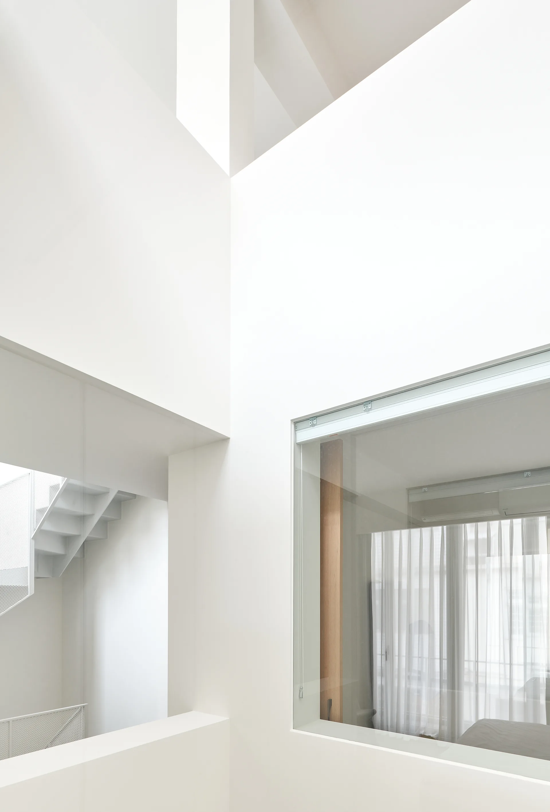A minimalist white multi-level interior features a metal staircase, geometric walls, and a large interior window revealing sheer curtains.