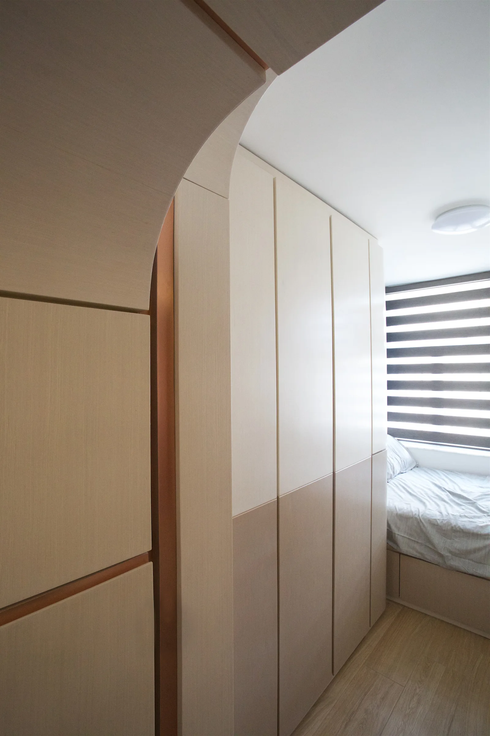 A minimalist bedroom features a custom light wood archway transition next to a floor-to-ceiling wardrobe and a bed by a window.