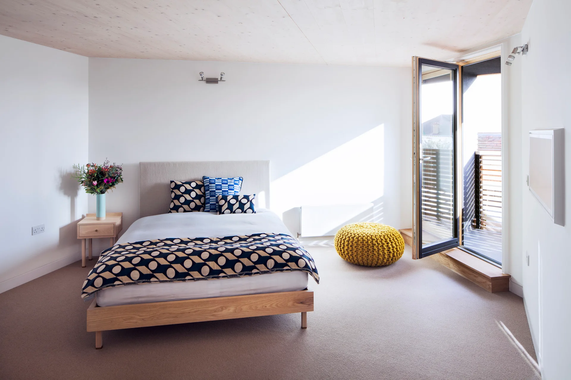A minimalist bedroom featuring a wooden bed with geometric bedding, a light wood plank ceiling, and a yellow knitted pouffe near an open balcony door.