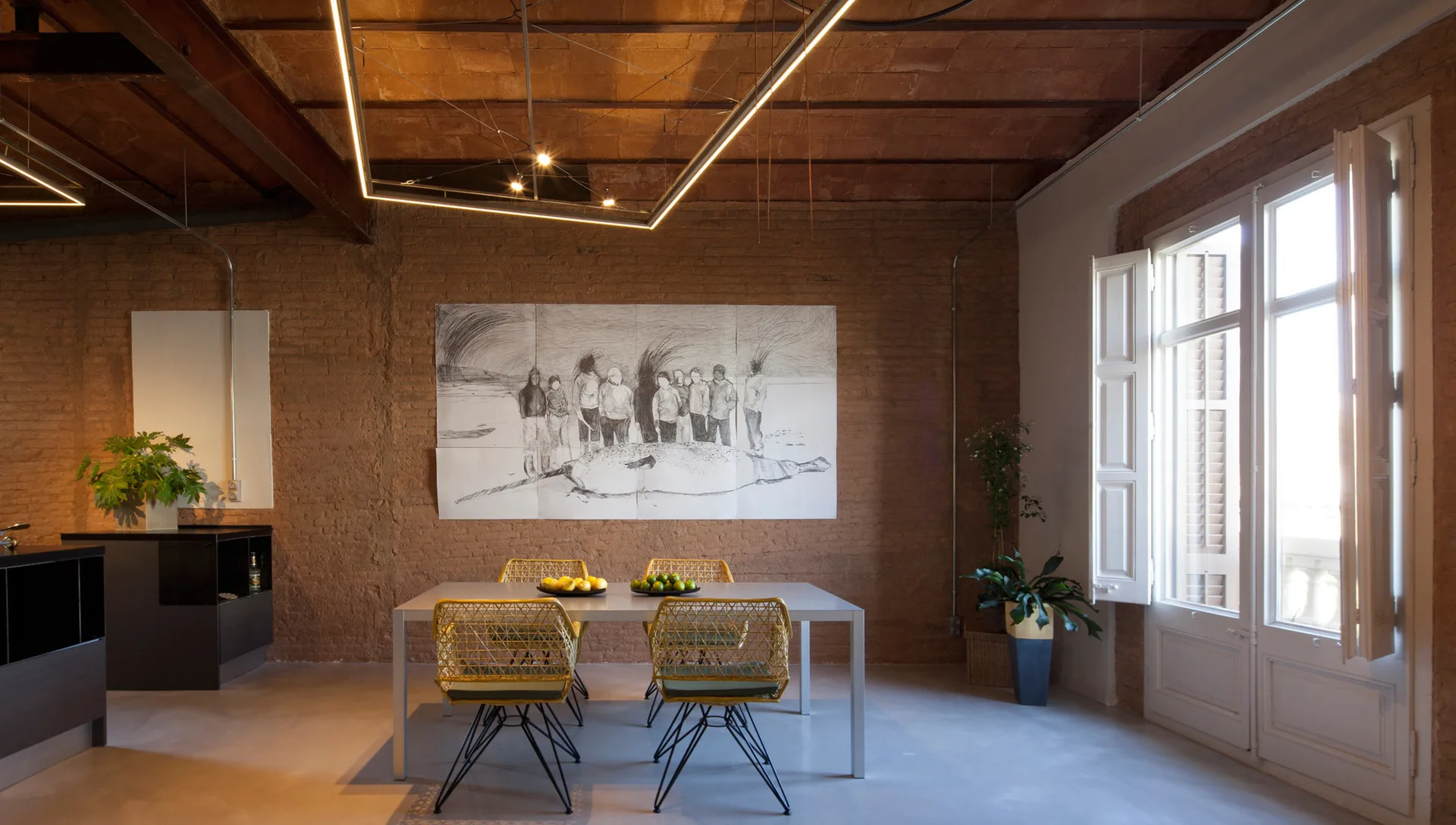 A grey dining table and yellow wire chairs sit under a vaulted brick ceiling with a large sketch hanging on the exposed brick wall.