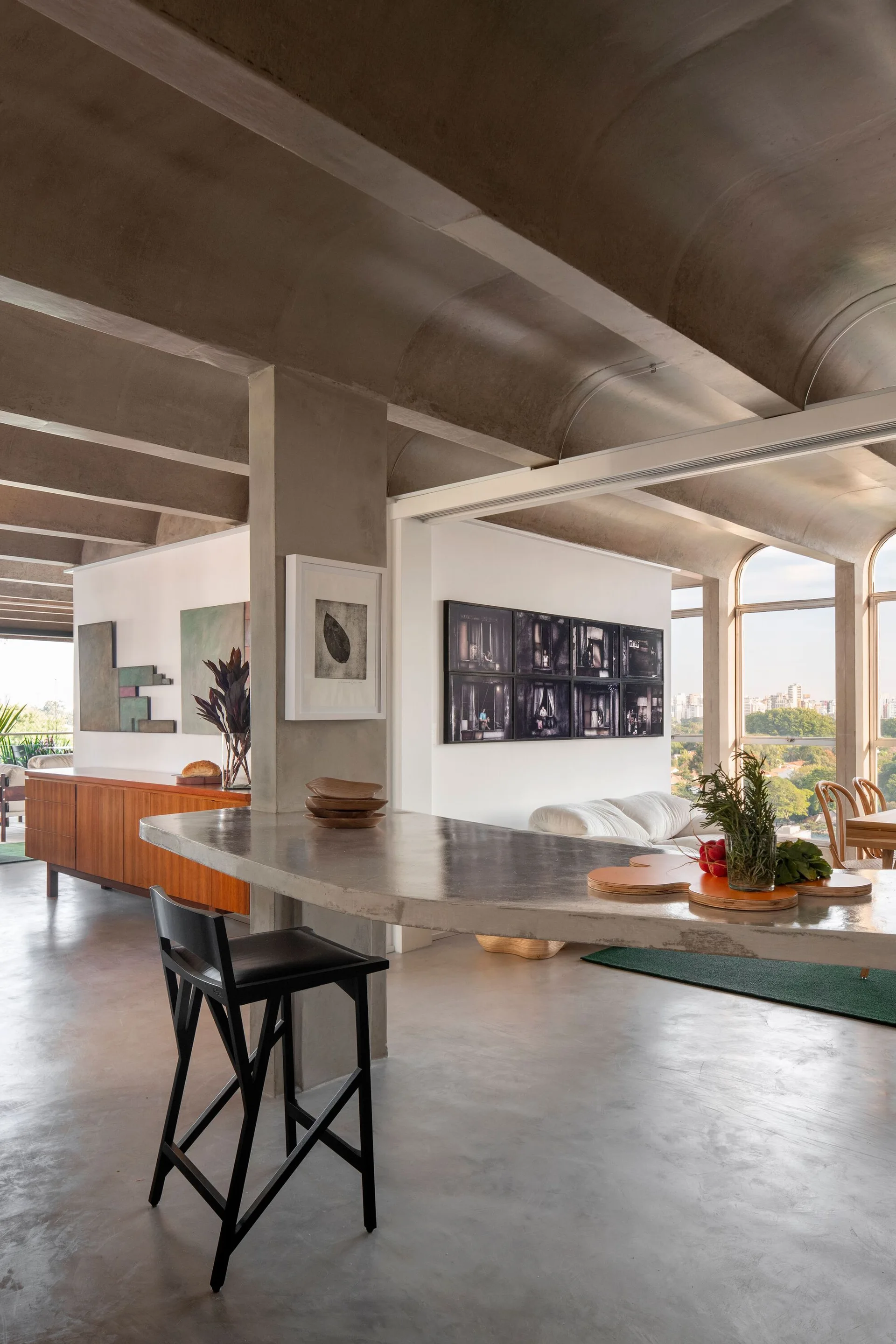 A modern open-plan interior featuring a concrete vaulted ceiling, a long cantilevered concrete island counter with a black bar stool, and a white sofa.