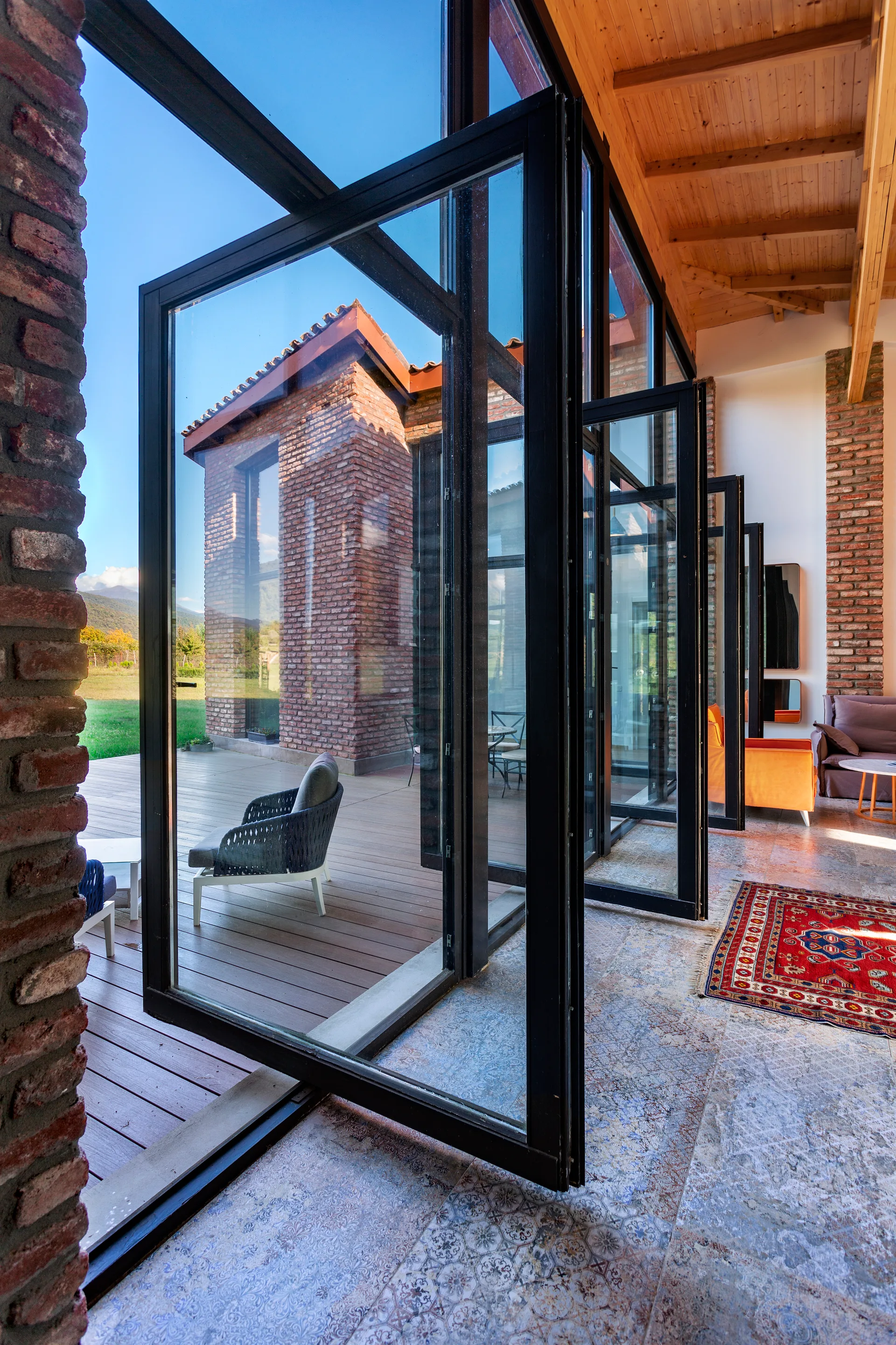 Large black-framed folding glass doors open from an indoor living area with exposed brick walls onto an outdoor wooden patio deck.