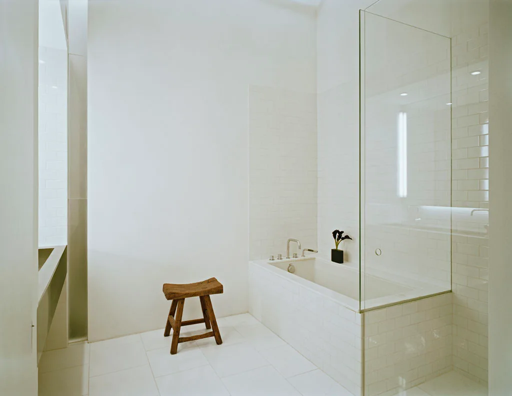 A minimalist white bathroom features a tiled built-in bathtub with a frameless glass partition and a rustic wooden stool on a tiled floor.