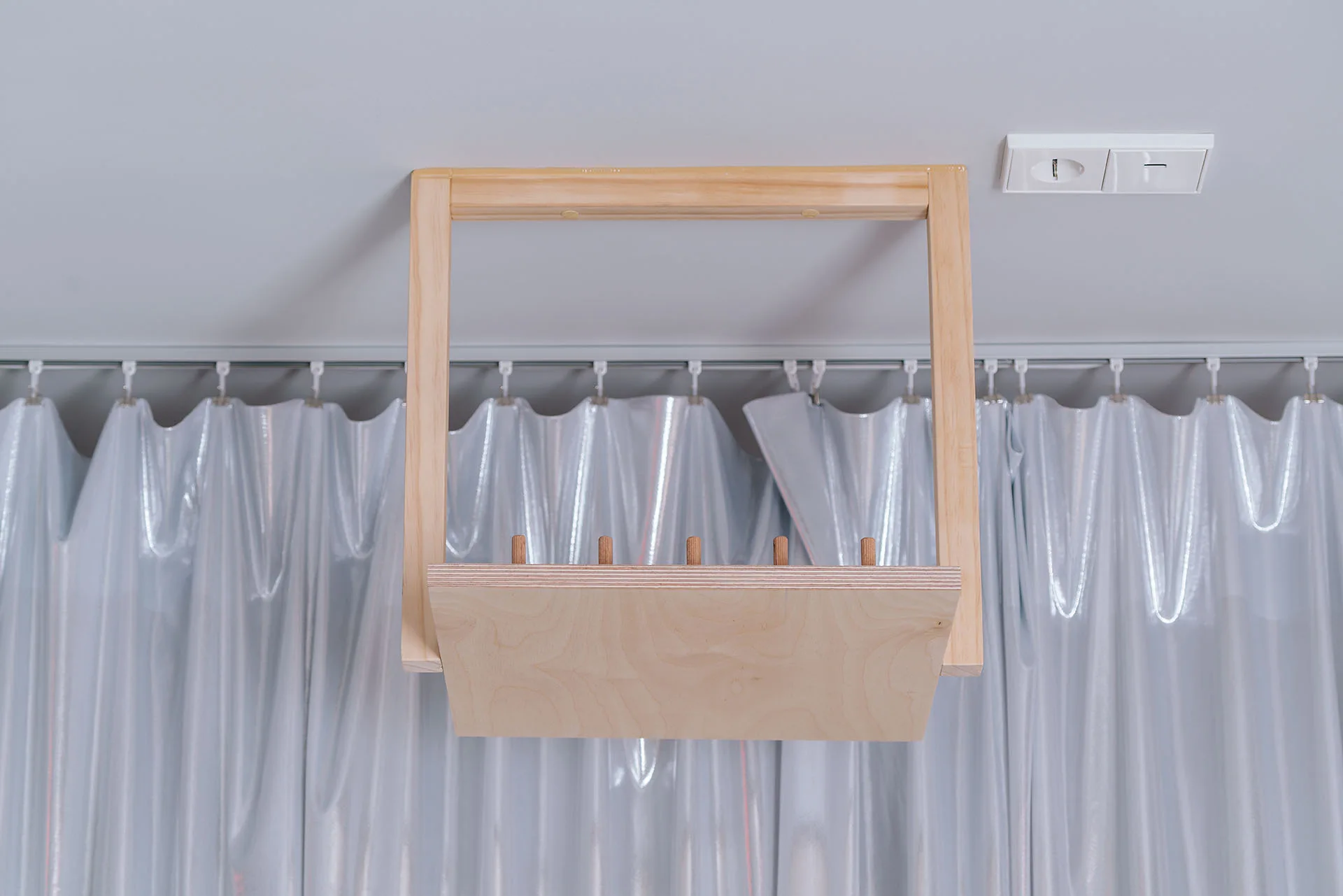 A rectangular light wood wall-mounted organizer with pegs hangs against a backdrop of draped light grey curtains.