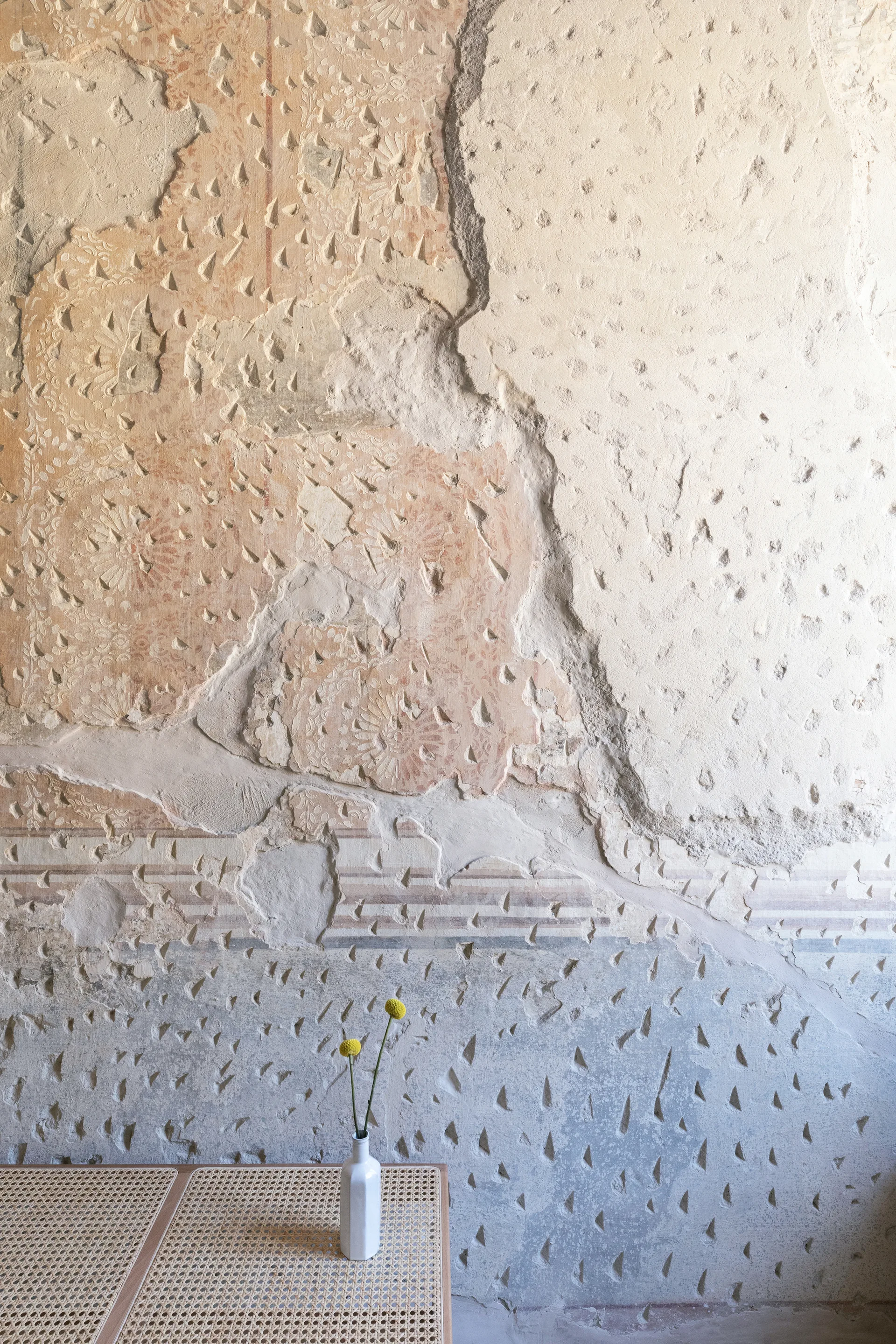 Two yellow flowers in a white ceramic vase rest on a woven cane tabletop in front of a heavily distressed, patterned plaster wall.