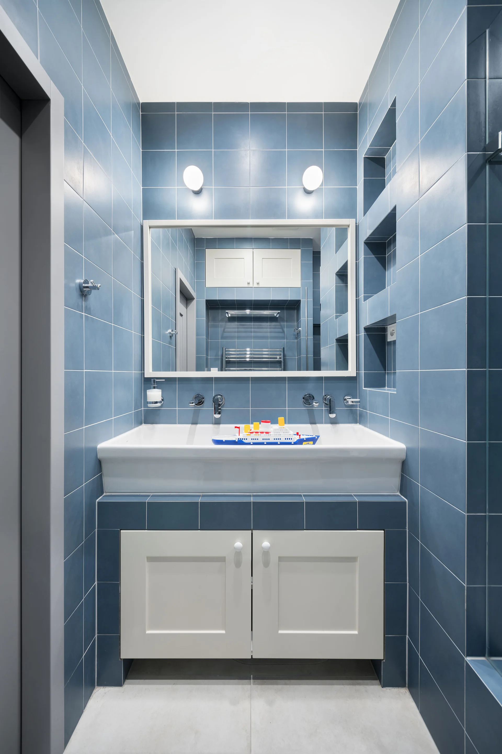 A modern bathroom vanity features blue tiled walls, a white ceramic sink, a rectangular mirror, and vertical recessed wall shelves.