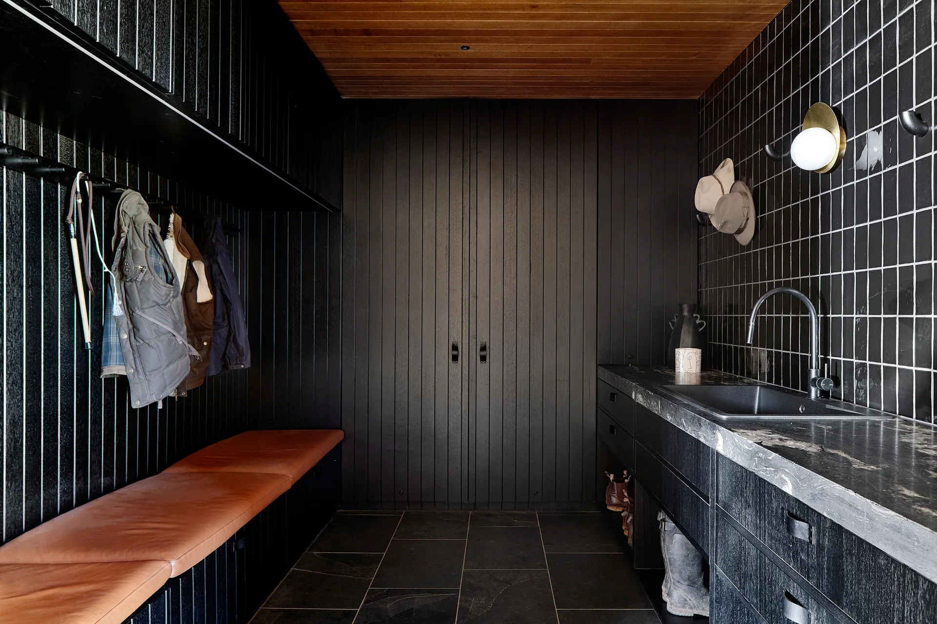 A dark mudroom featuring vertical black wood paneling, a tan leather bench, a dark stone vanity with a sink, and a warm wood plank ceiling.