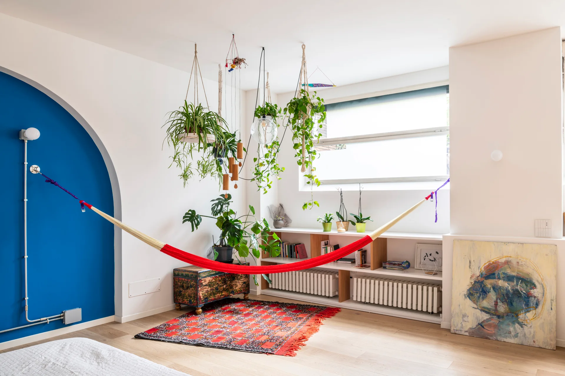A bright living room featuring a suspended red hammock, a painted blue wall arch, hanging potted plants, and a low white bookshelf.
