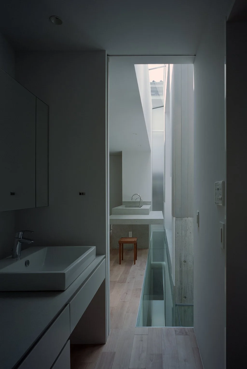 A minimalist bathroom corridor features a white vanity, a glass floor strip beside an exposed concrete column, and a skylight overhead.