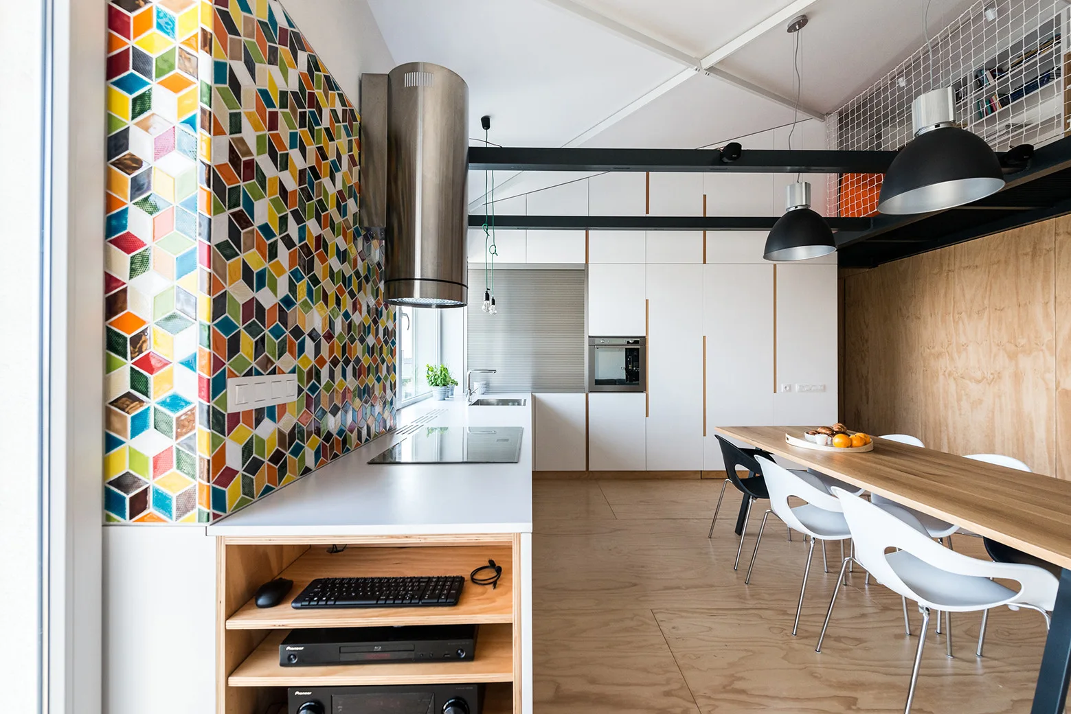A modern kitchen featuring a colorful geometric patterned tile wall, white cabinetry, a light wood dining table, and black ceiling beams.