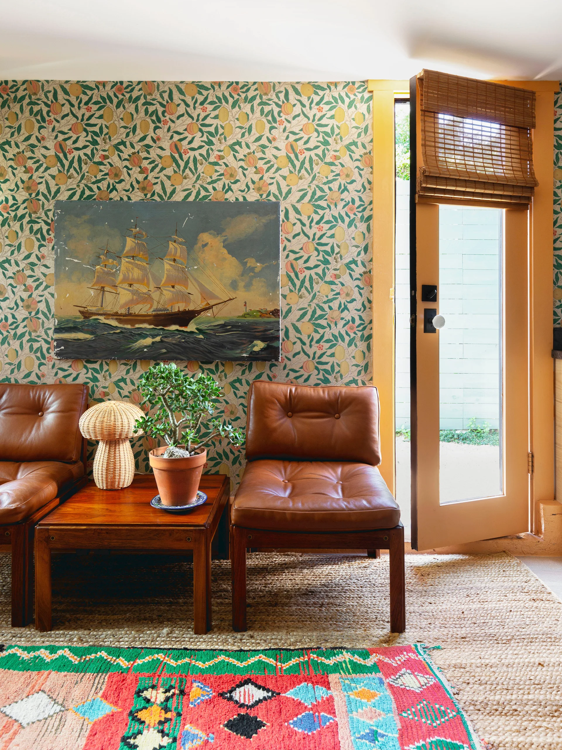 Two brown tufted leather chairs stand beside a wooden table with a mushroom lamp, set against botanical wallpaper next to an open door.