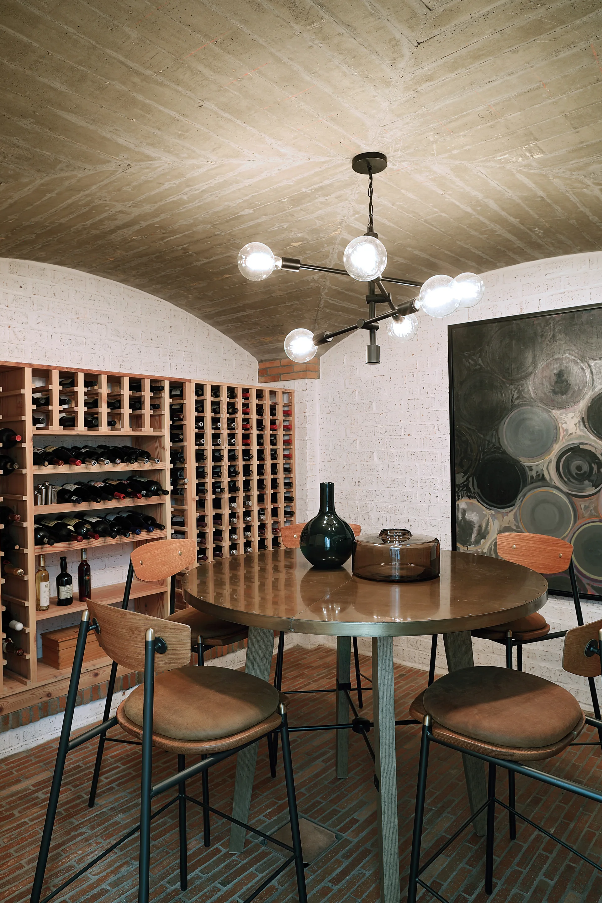 A round metal table with chairs sits beneath a modern geometric chandelier in a wine cellar with brick walls and a vaulted concrete ceiling.