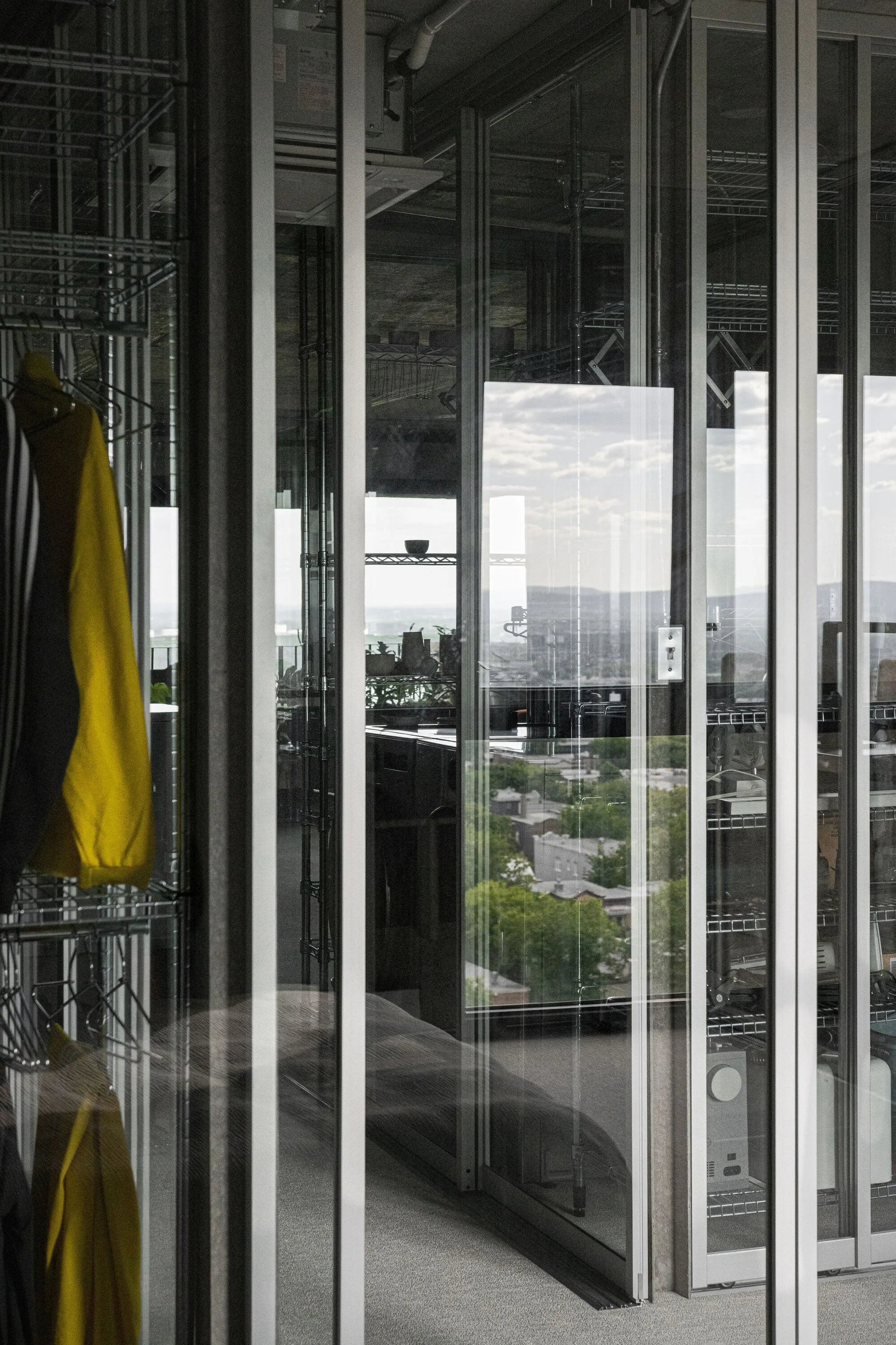 A modern interior features metal-framed glass partitions reflecting an urban view next to industrial wire shelving units.