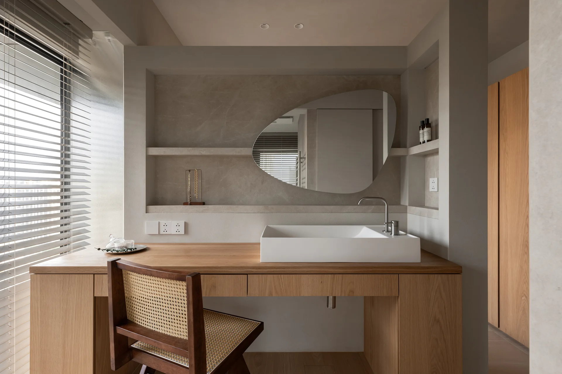 A wooden bathroom vanity with a white vessel sink, an organic-shaped mirror, and a cane chair set against a recessed stone-textured niche.