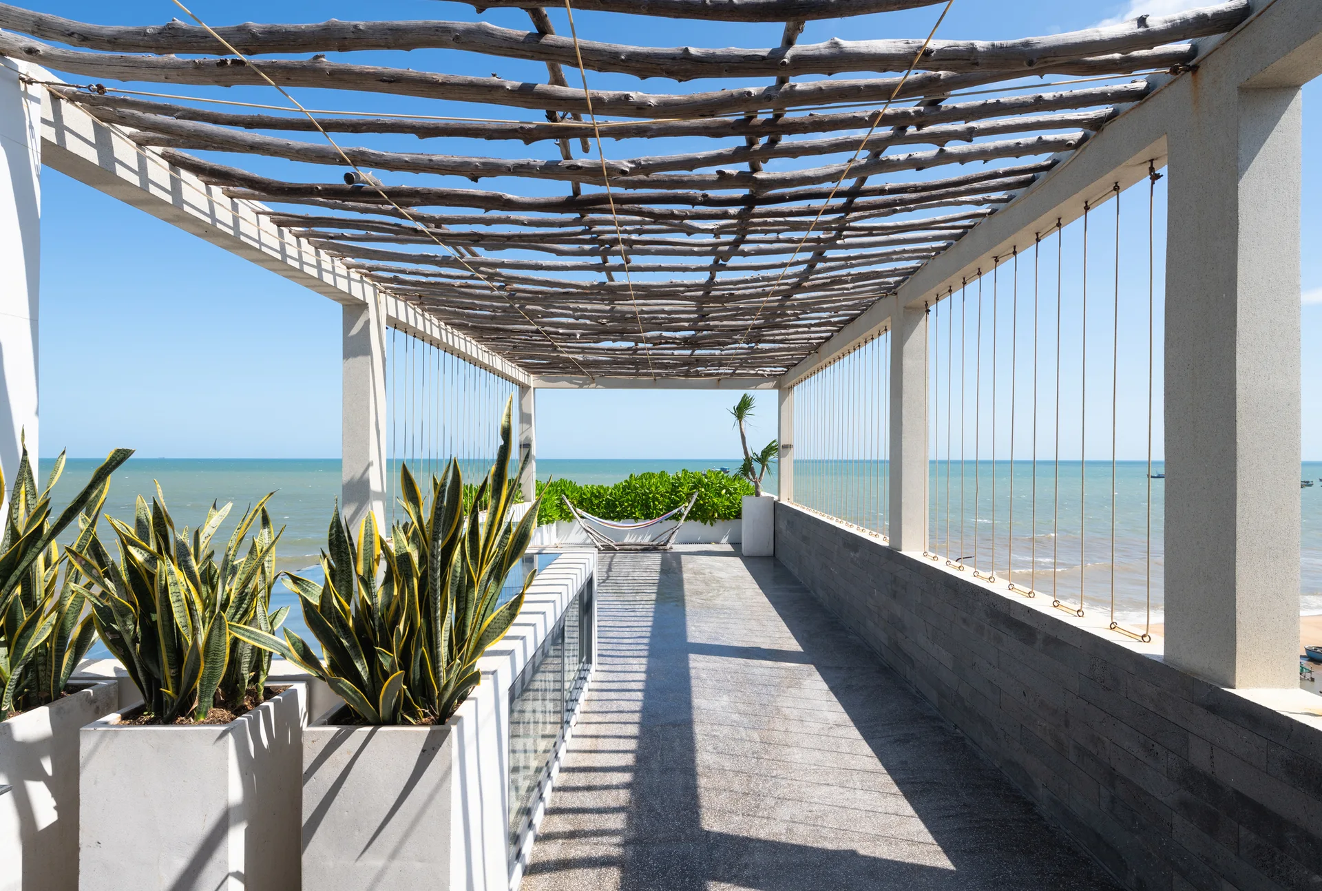 A sunlit coastal terrace features a rustic branch pergola overhead, a low stone wall with rope railing, and snake plants in white rectangular planters.