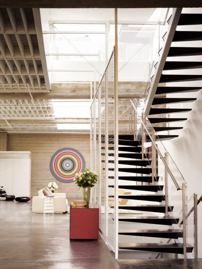 An industrial loft features a black floating metal staircase next to a white mesh partition, a polished concrete floor, and a concrete waffle ceiling.